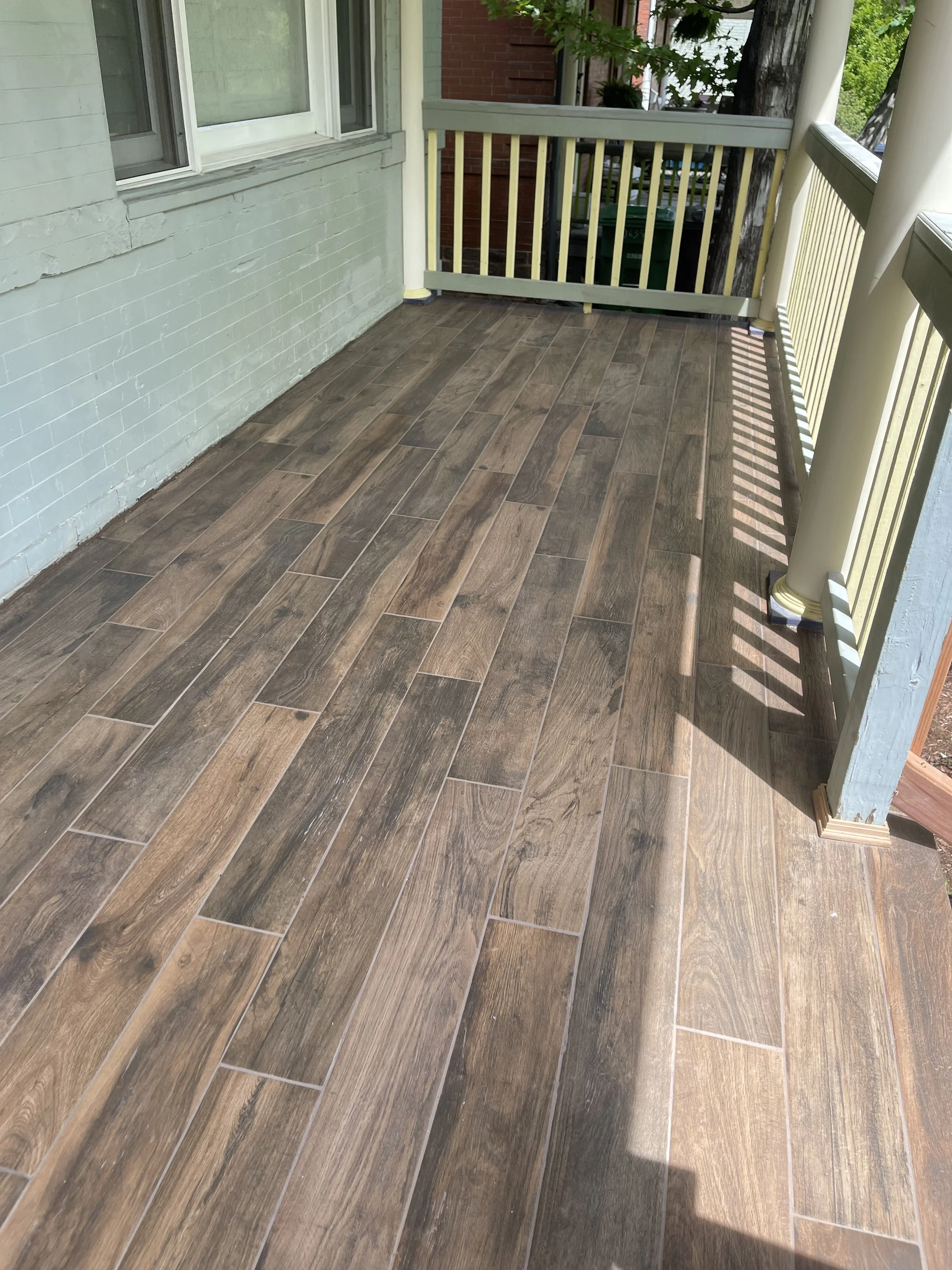 Covered porch renovation by Otto Custom Builders featuring wood-look tile flooring, a painted brick wall, windows, and yellow-and-gray railings. Professional porch remodeling, tile flooring installation, exterior painting, and home improvement.