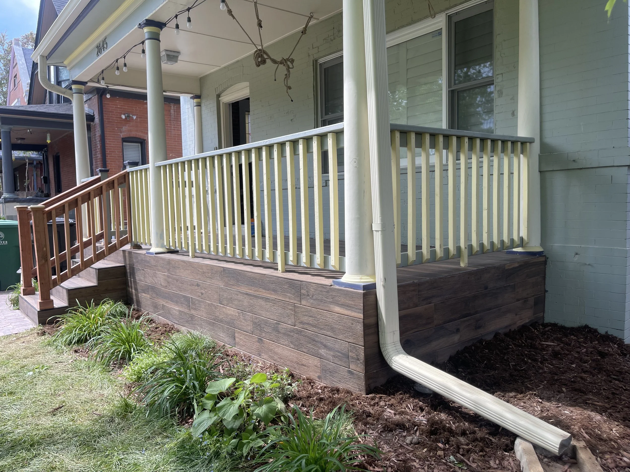 Exterior home renovation by Otto Custom Builders featuring a blue brick front porch, white columns, pale yellow railings, wooden steps, string lights, and landscaped garden beds. Professional porch remodeling, exterior painting, wood repair, and home