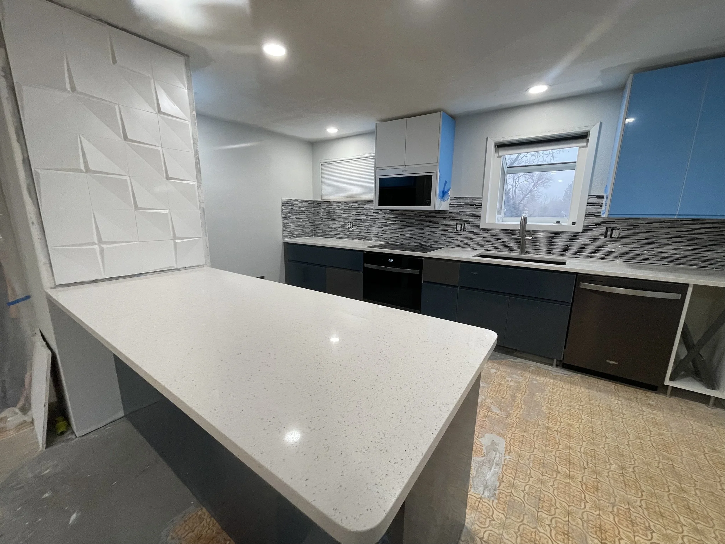 Modern kitchen remodeling by Otto Custom Builders featuring a large white island countertop, blue and white kitchen cabinets, stainless steel appliances, a sink, and a mosaic tile backsplash. Professional kitchen renovation, cabinet installation.