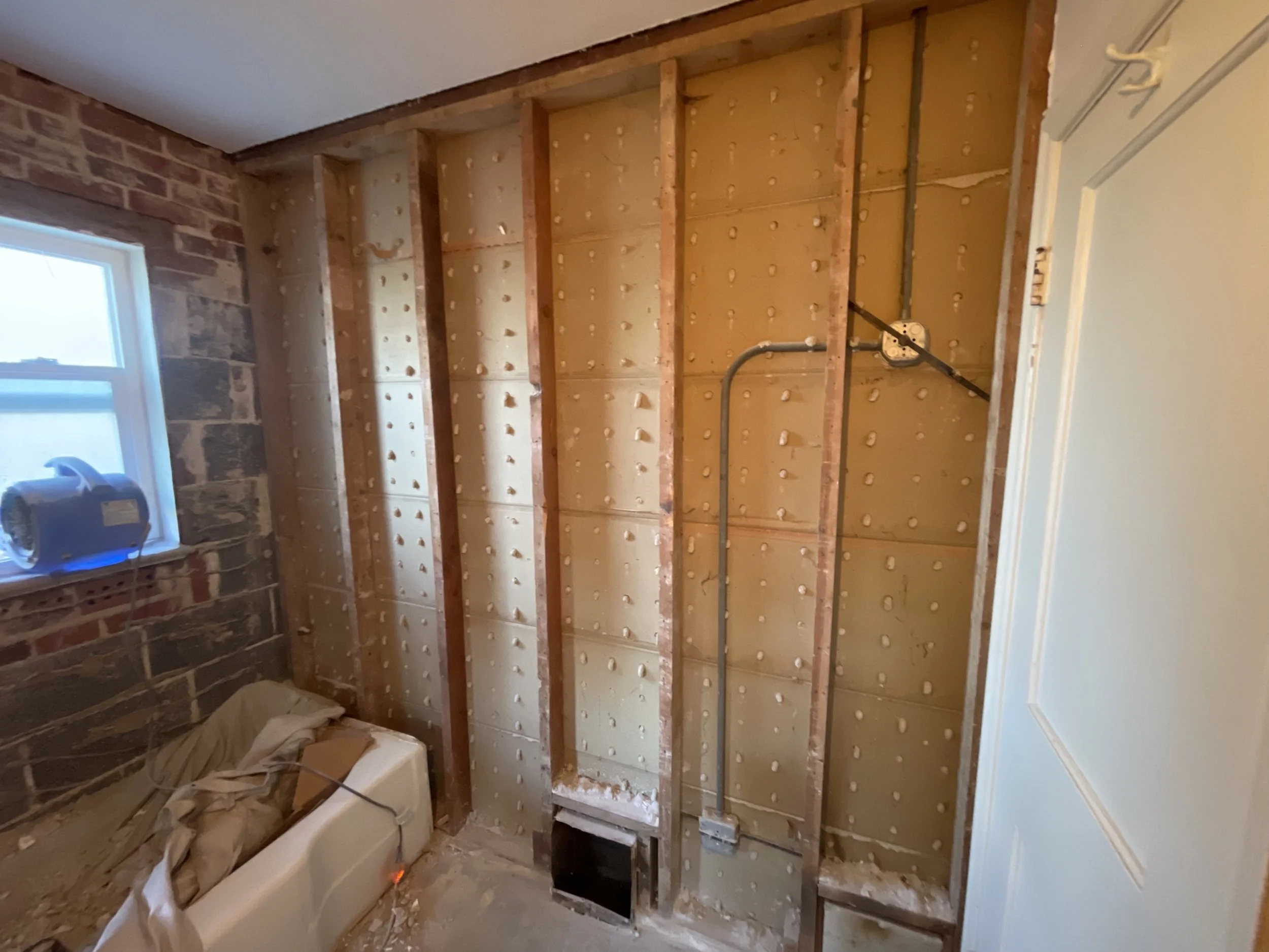 Residential bathroom demolition and remodeling by Otto Custom Builders, featuring exposed wood wall studs, insulation, electrical wiring, brickwork, a window, and bathtub in the Denver Metro area, Colorado