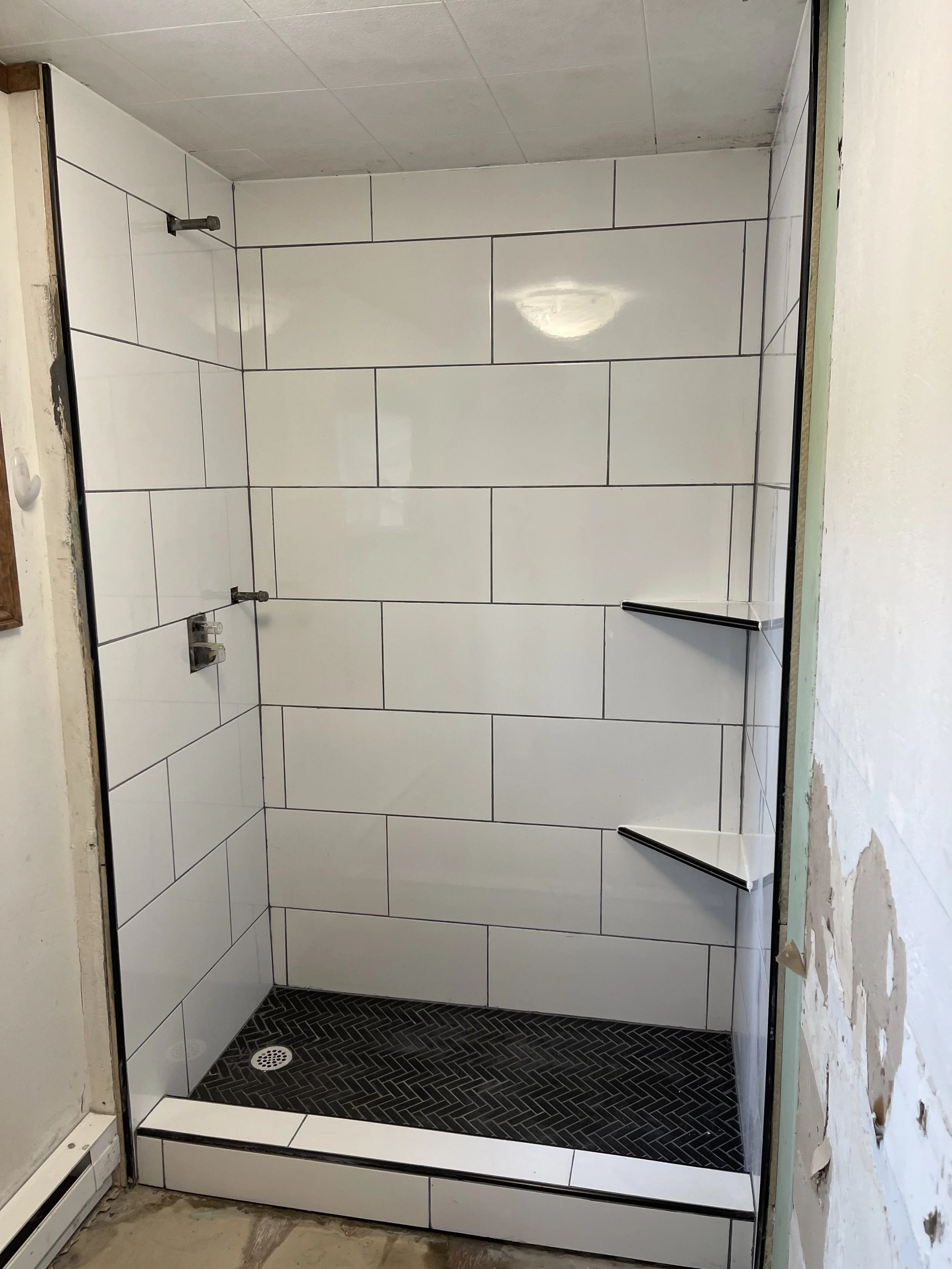 Walk-in shower remodeling by Otto Custom Builders featuring white subway tile walls, black herringbone tile flooring, modern shower fixtures, a built-in shower bench, and a floor drain. Professional bathroom remodeling, shower tile installation.