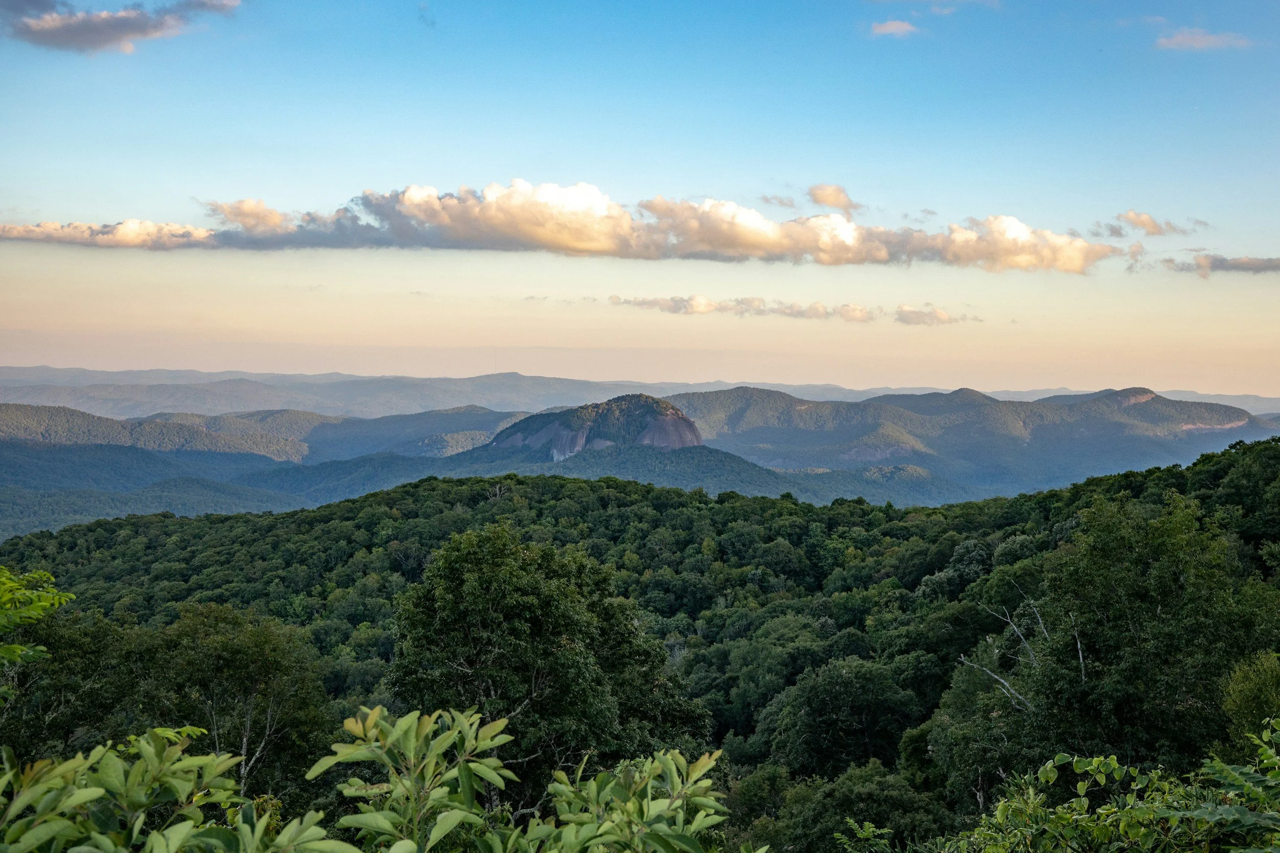 Scenic view of a lush green forested mountain range with layers of mountains in the distance and a partly cloudy sky overhead.
