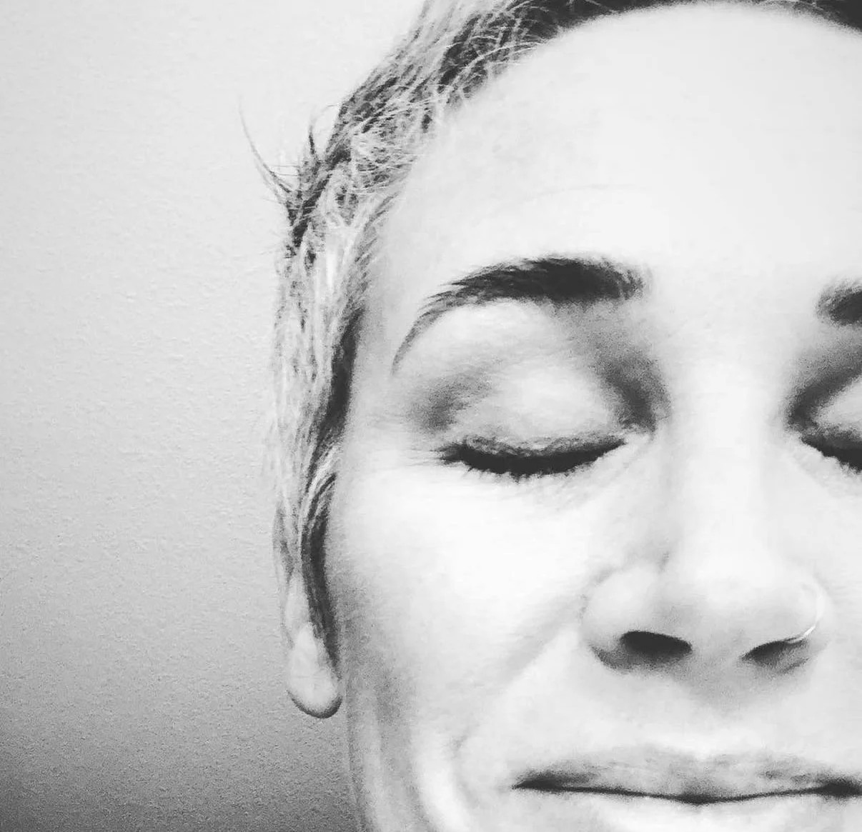 Close-up black-and-white photo of a woman with closed eyes, light hair, and a nose piercing.