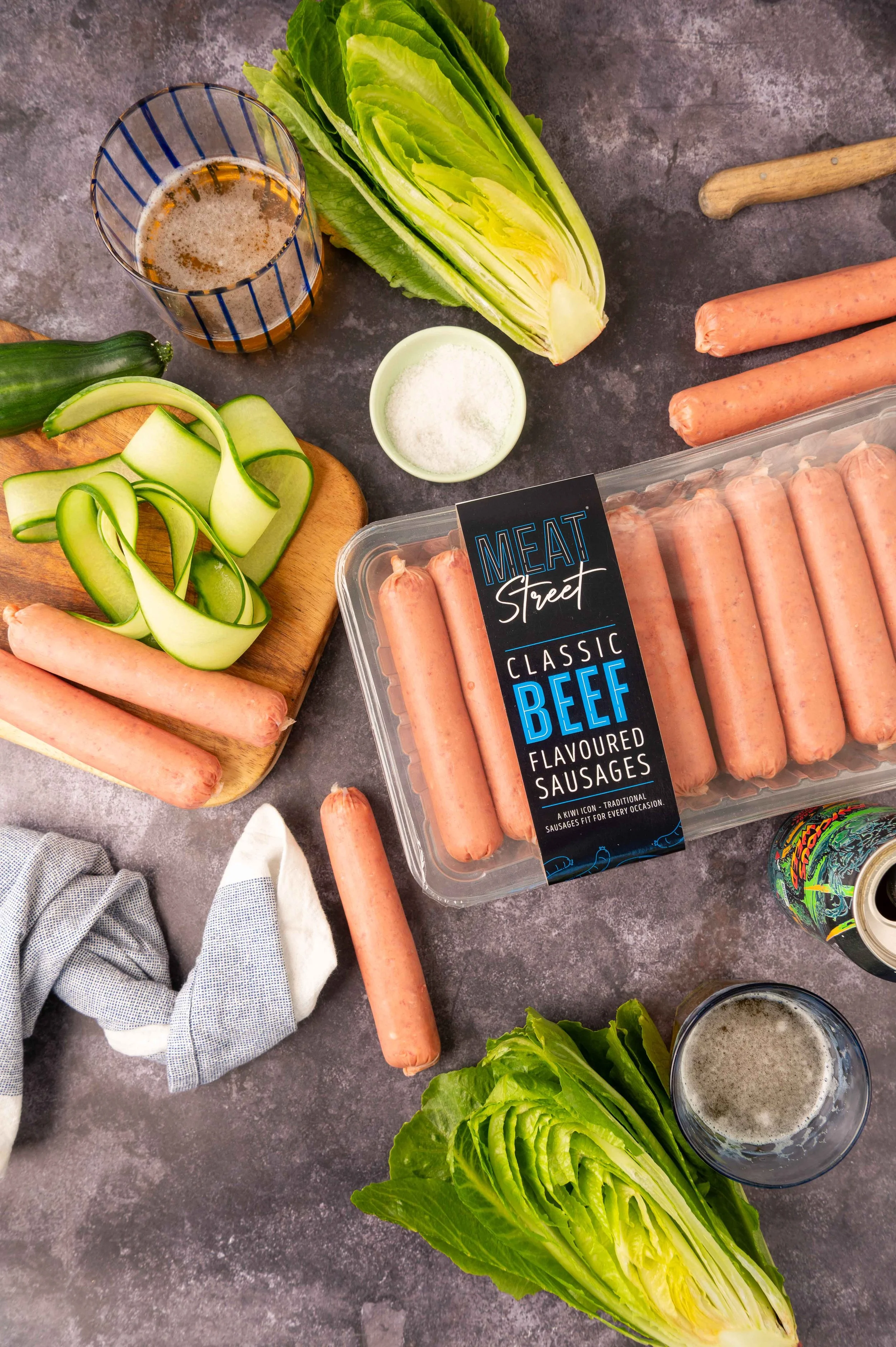 Meat Street Classic Sausages, shot surrounded by fresh lettuce, zucchini, carrots, a glass of beer, and a can of soda on a dark countertop.
