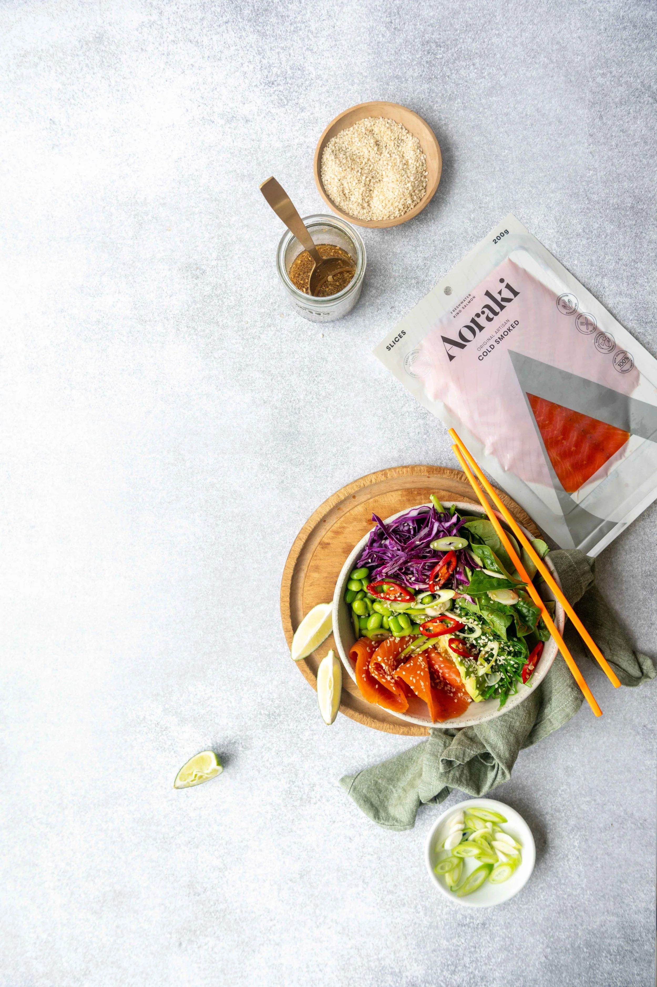 A salad with Aoraki Smoked Salmon and Japanese Salad sits on a Chopping Borad. Lime wedges and a small bowl of sliced green onions sit nearby. A glass jar holds Japanese Dressing.