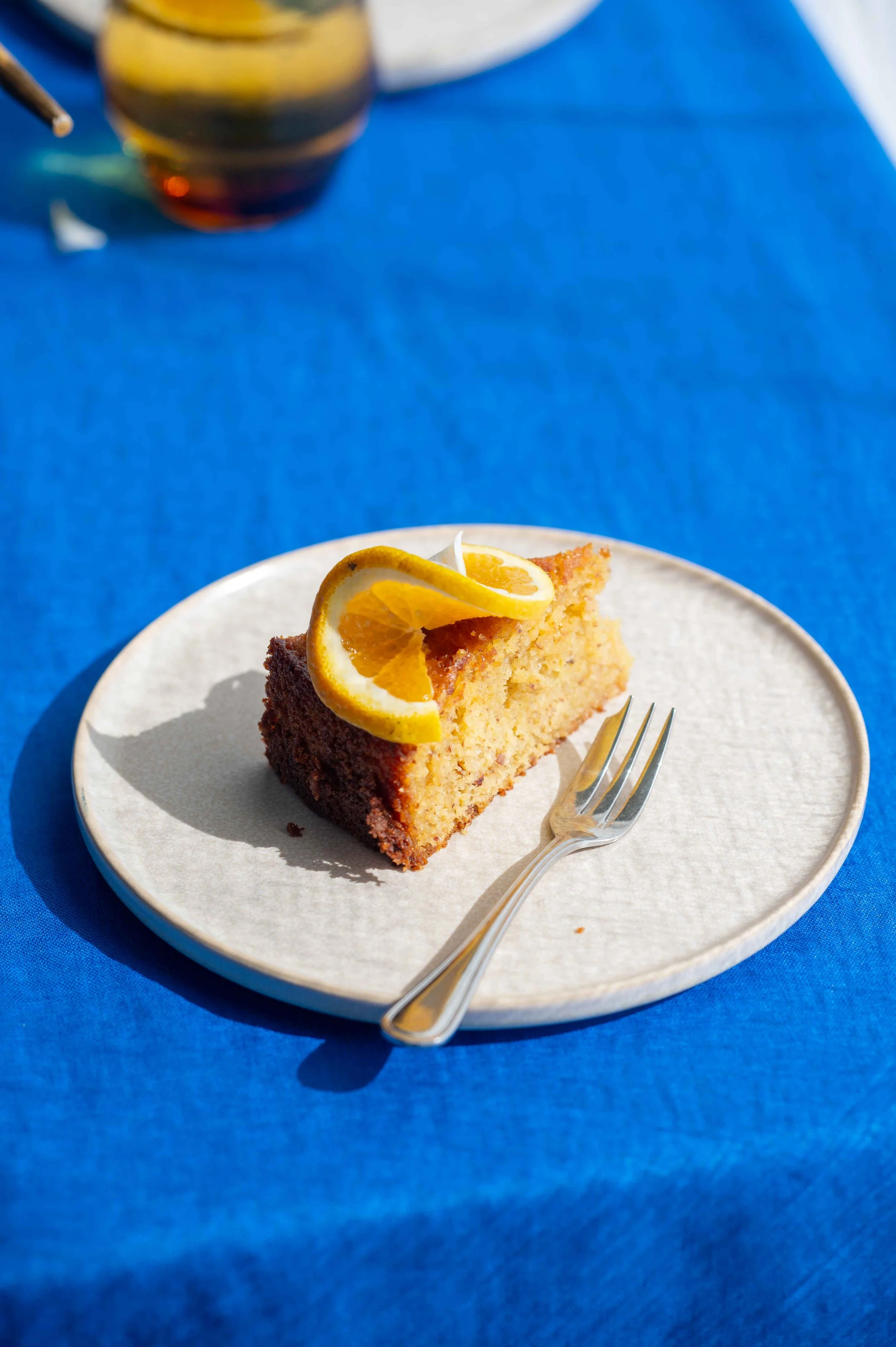 A slice of cake garnished with an orange twist. Water in an amber glass. Shot on blue linen, natural light.