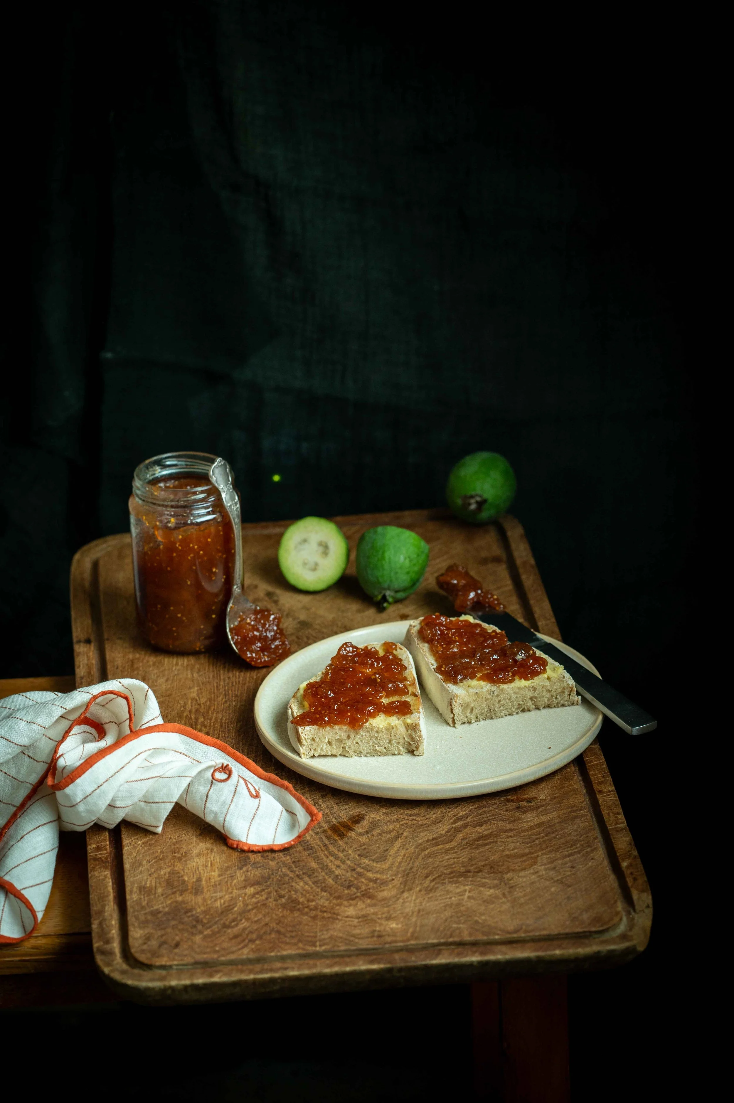 A jar of Feijoa & Ginger Jam sits on a wooden chopping board next to a white plate with sourdough bread topped with butter and jam. Shot dark and moody.