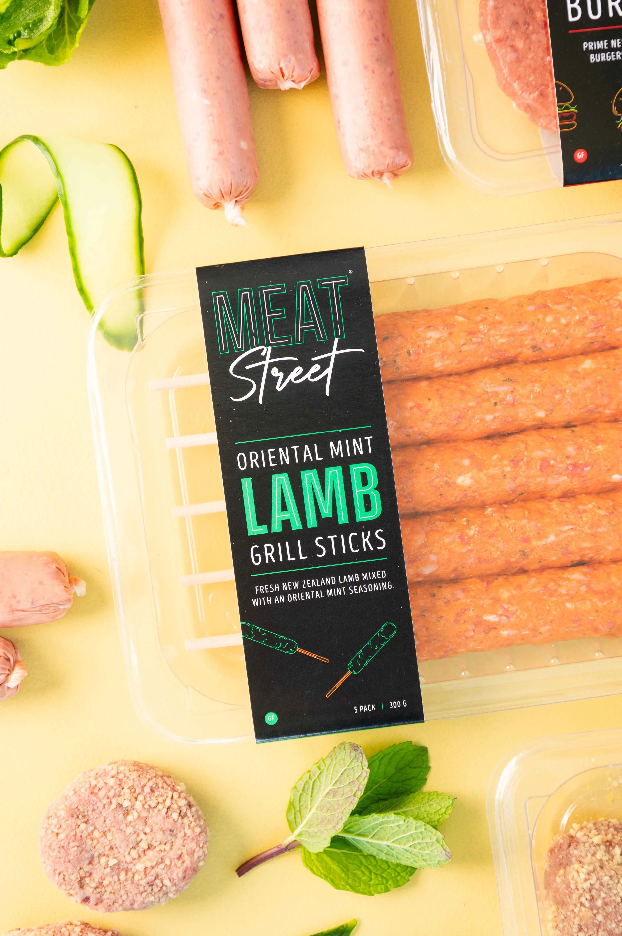 Package of Meat Street Oriental Mint Lamb Grill Sticks with fresh lamb, cucumber slices, mint leaves, and other food items on a yellow background.