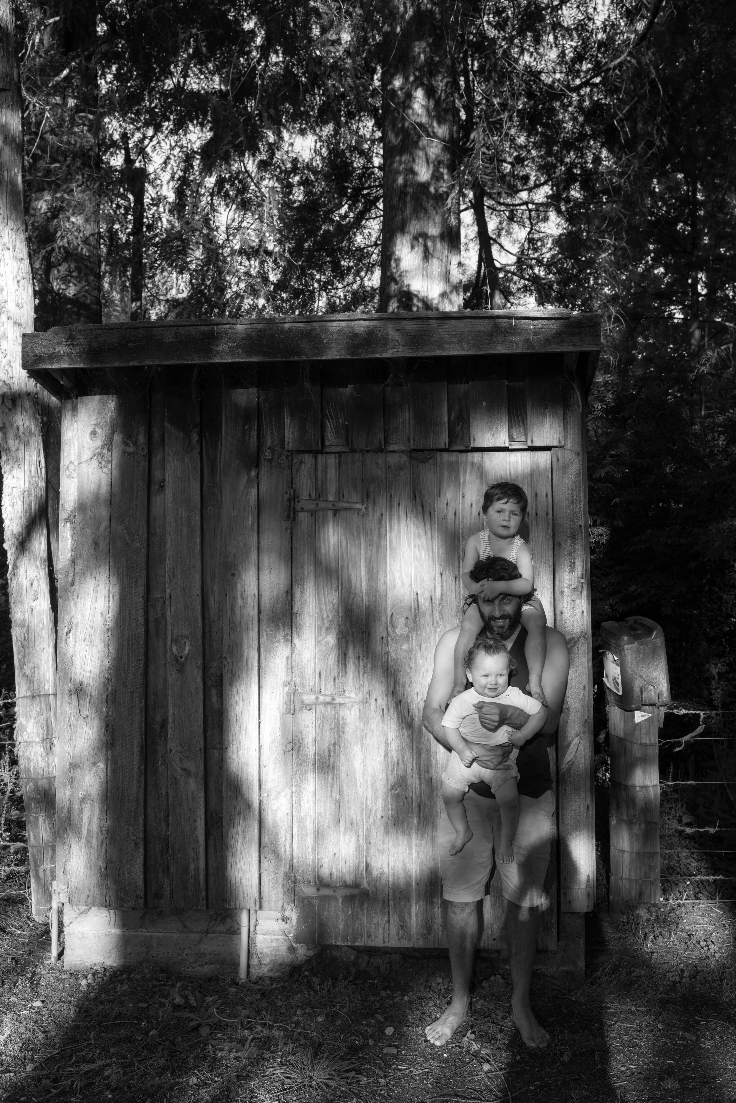 A man with two children standing and sitting in front of a wooden shed, in a forested outdoor area, with shadows of trees cast on the shed and ground, black and white photo.