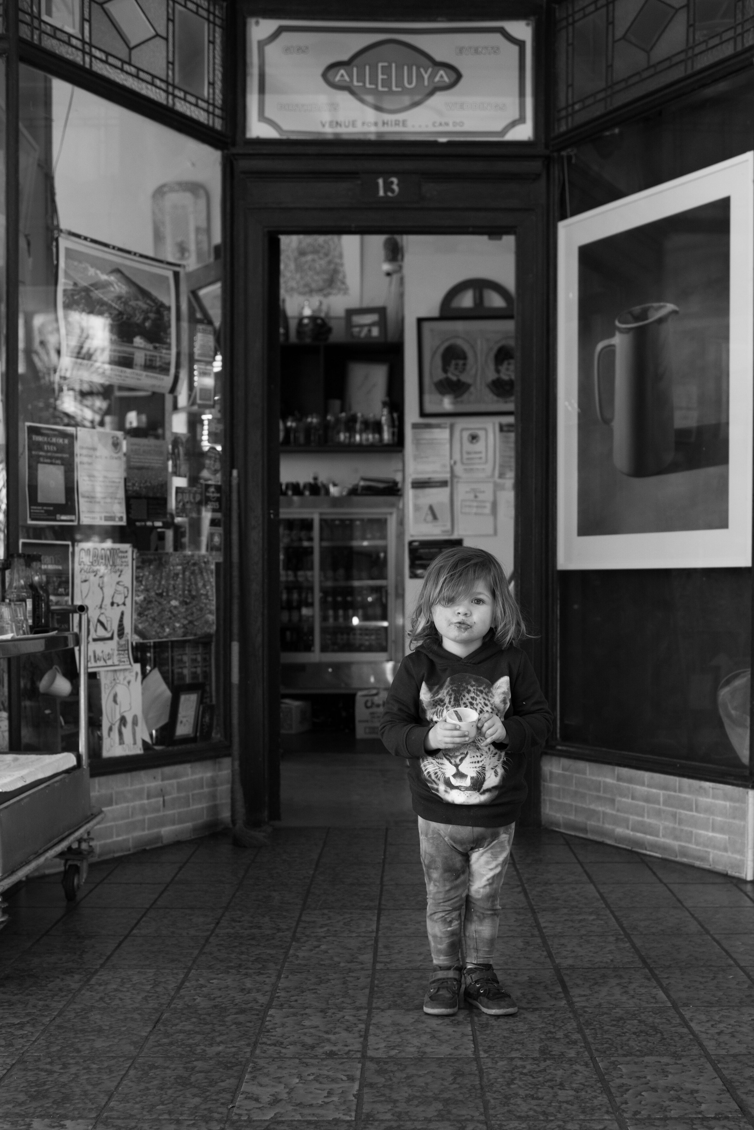 A young child with tousled hair standing inside a shop, holding a cup and making a duck face at the camera, with shelves, posters, and a large picture of a mug on the wall behind.