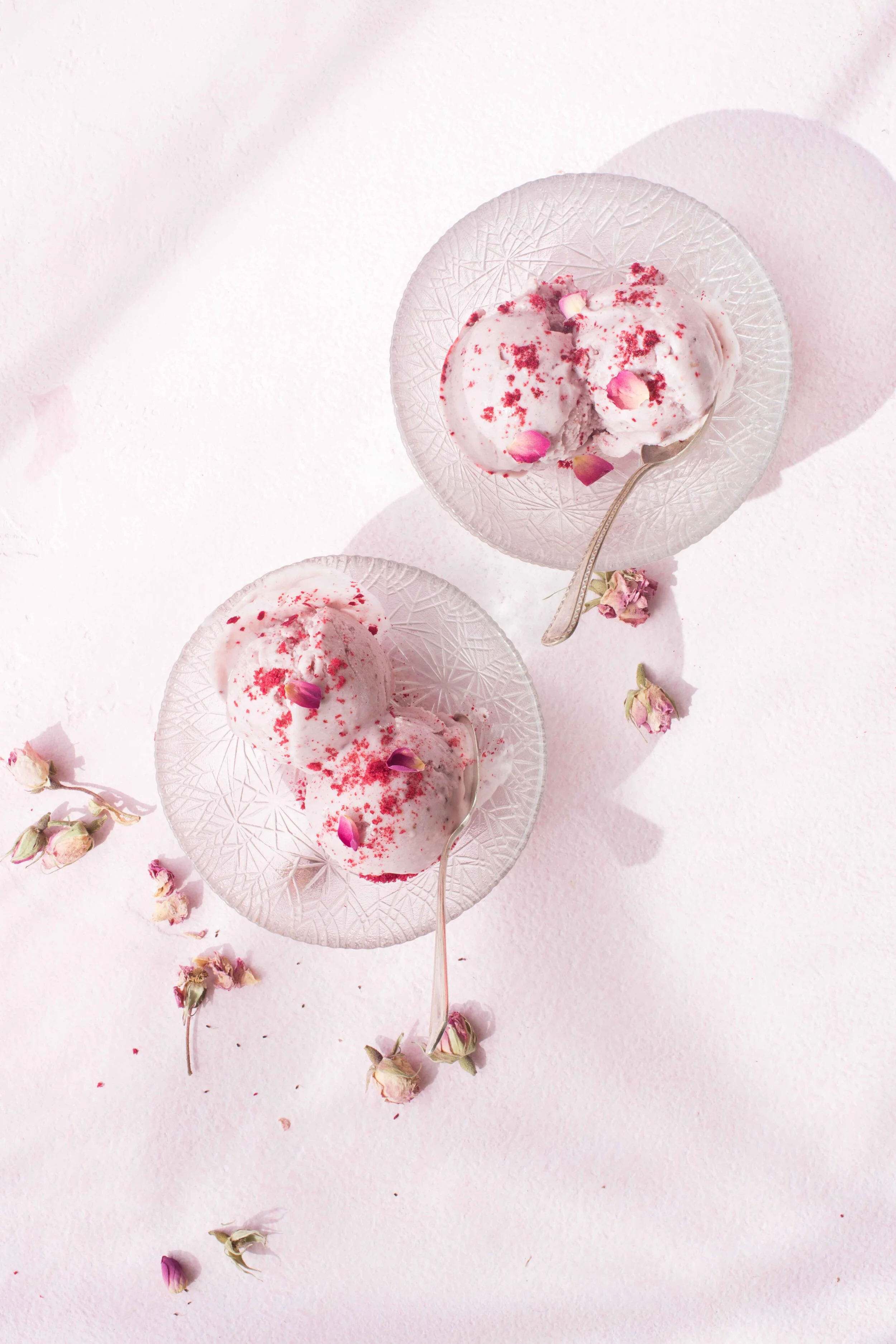Two glass bowls with scoops of pink ice cream, garnished with rose petals, on a light pink surface with scattered rosebuds.