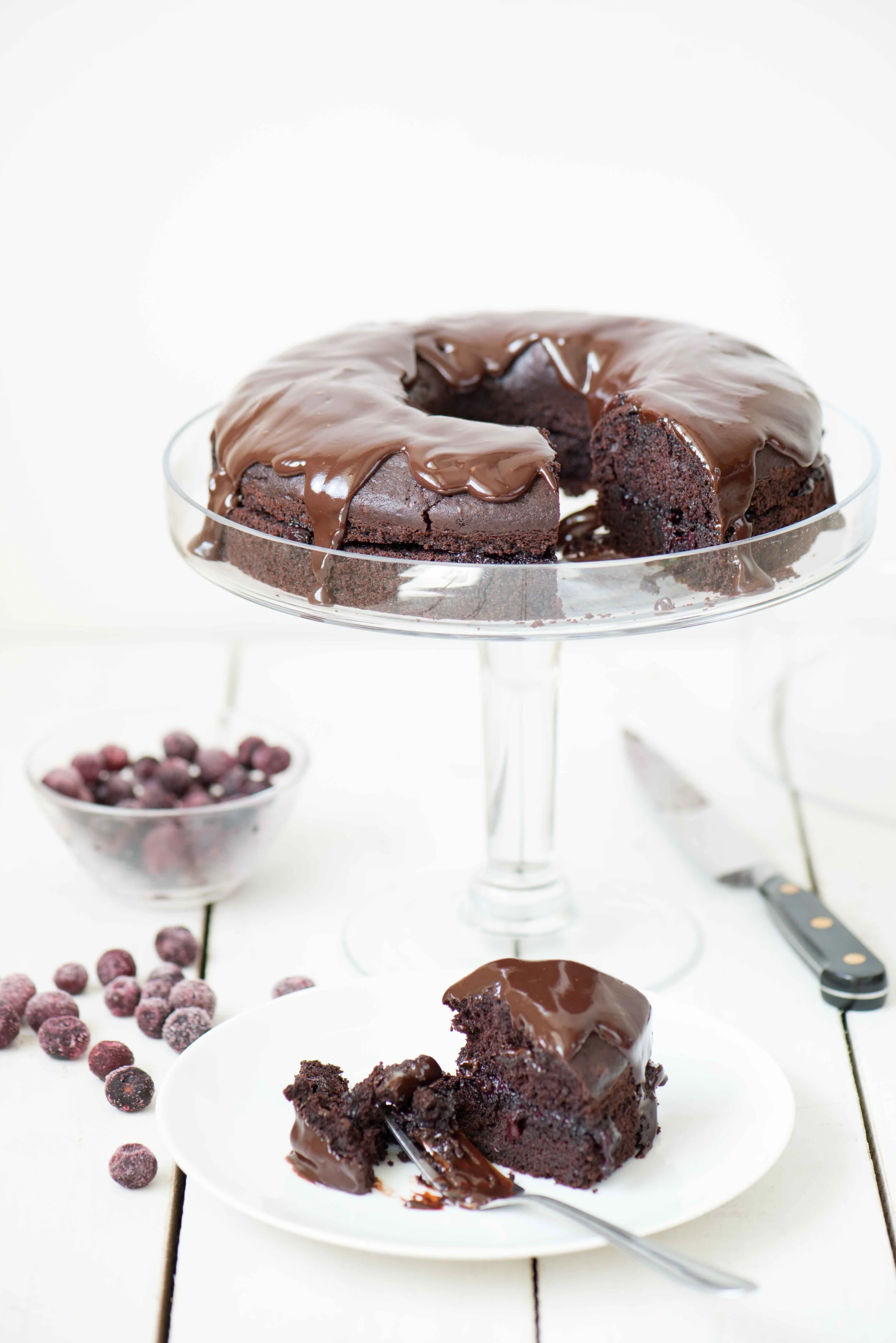 Chocolate cake with glossy chocolate glaze on a glass cake stand, with a slice served on a white plate, surrounded by blueberries and a knife.