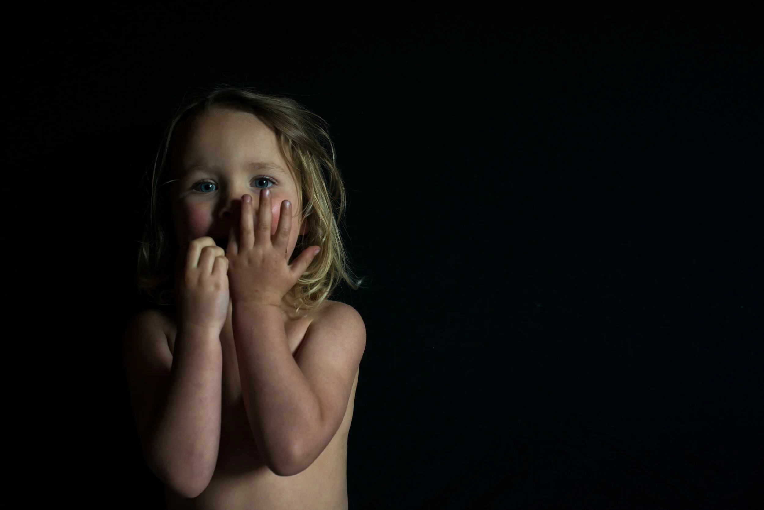 A young child with blonde hair and blue eyes covering their mouth with both hands against a dark background.