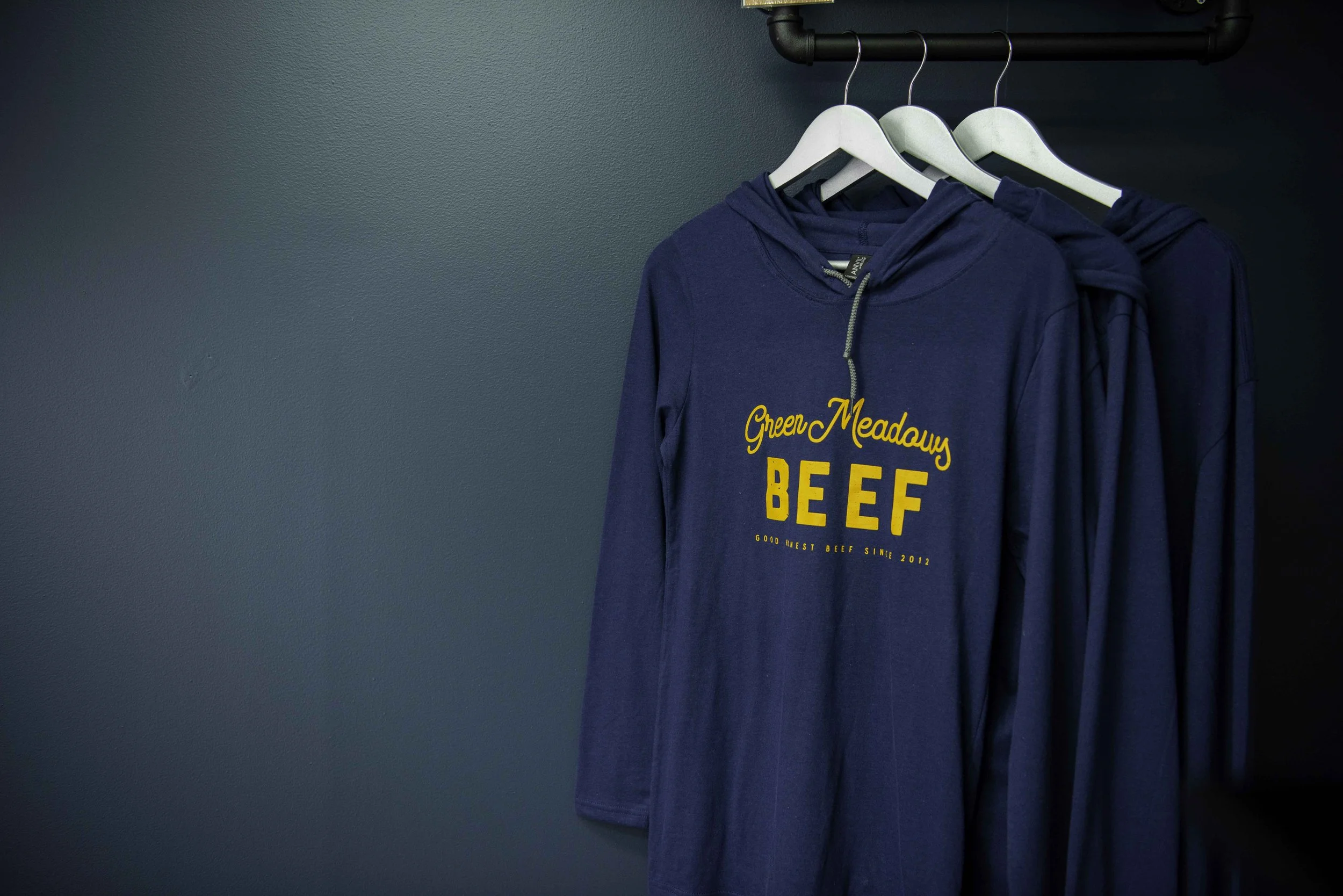 A Green Meadow’s Beef long sleeved hooded t-shirt handing on a rack. Shot against a dark wall.