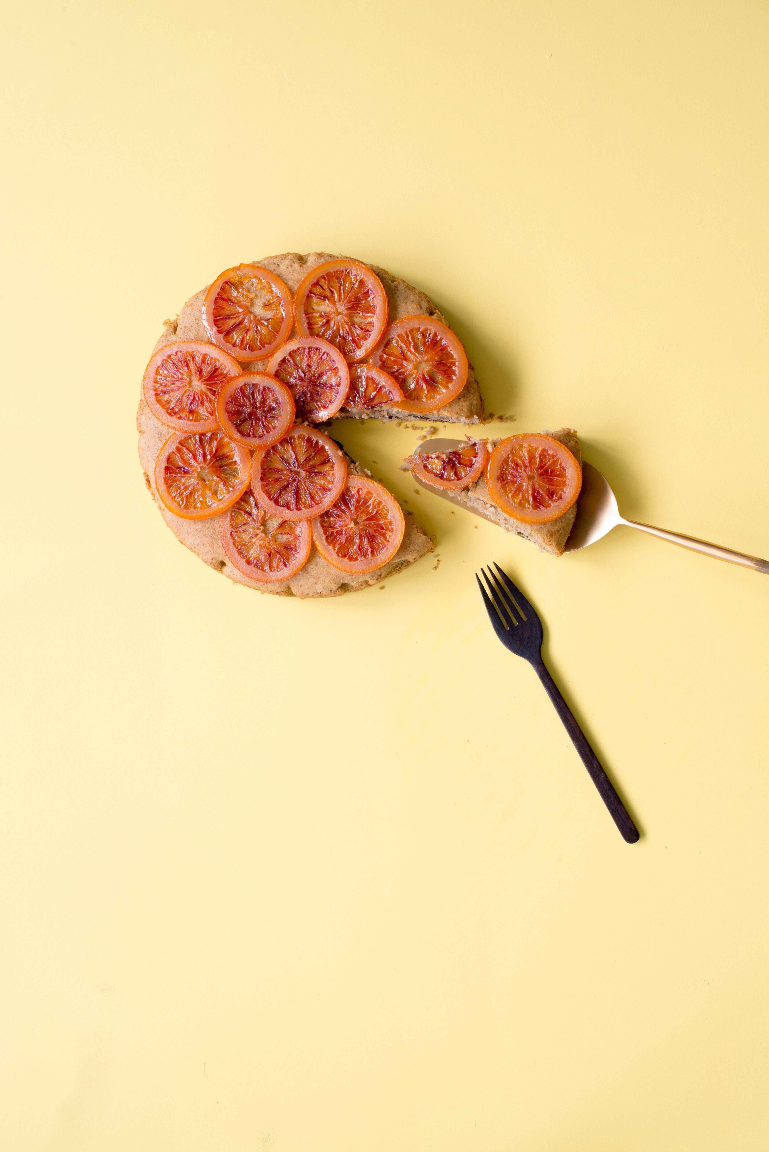 A sliced cake topped with blood orange slices on a yellow background, with a piece being cut and served on a fork.