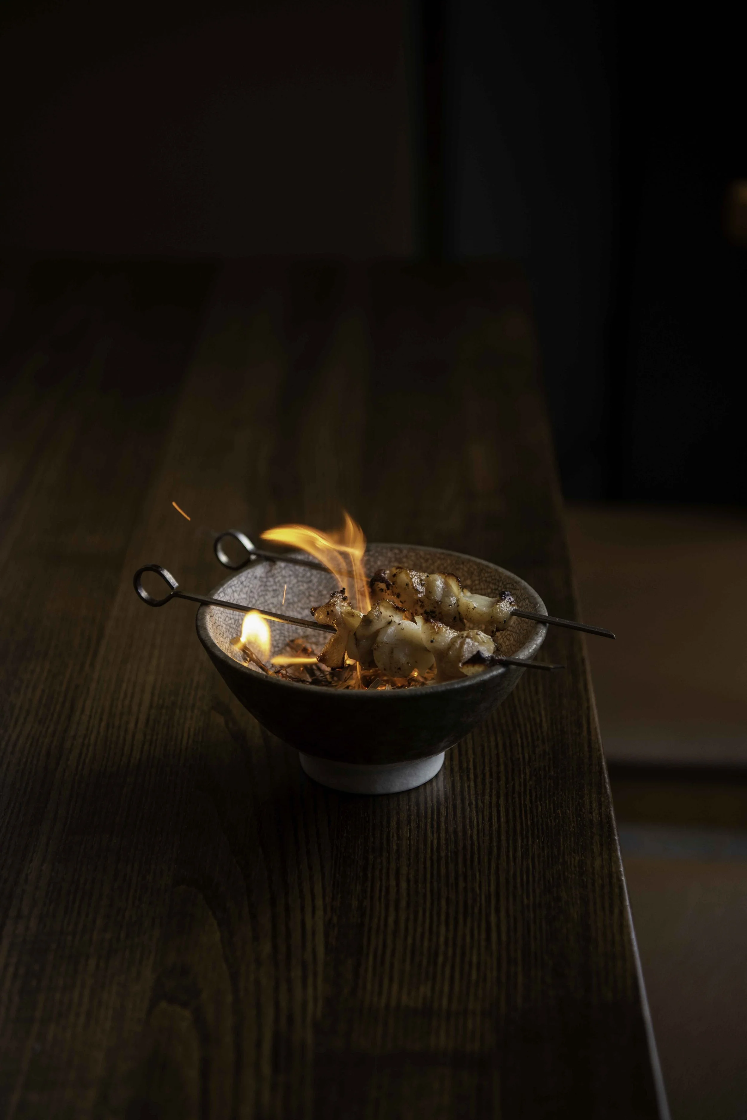A bowl with flaming skewers sits on a wooden dining table. Ember Restaurant.