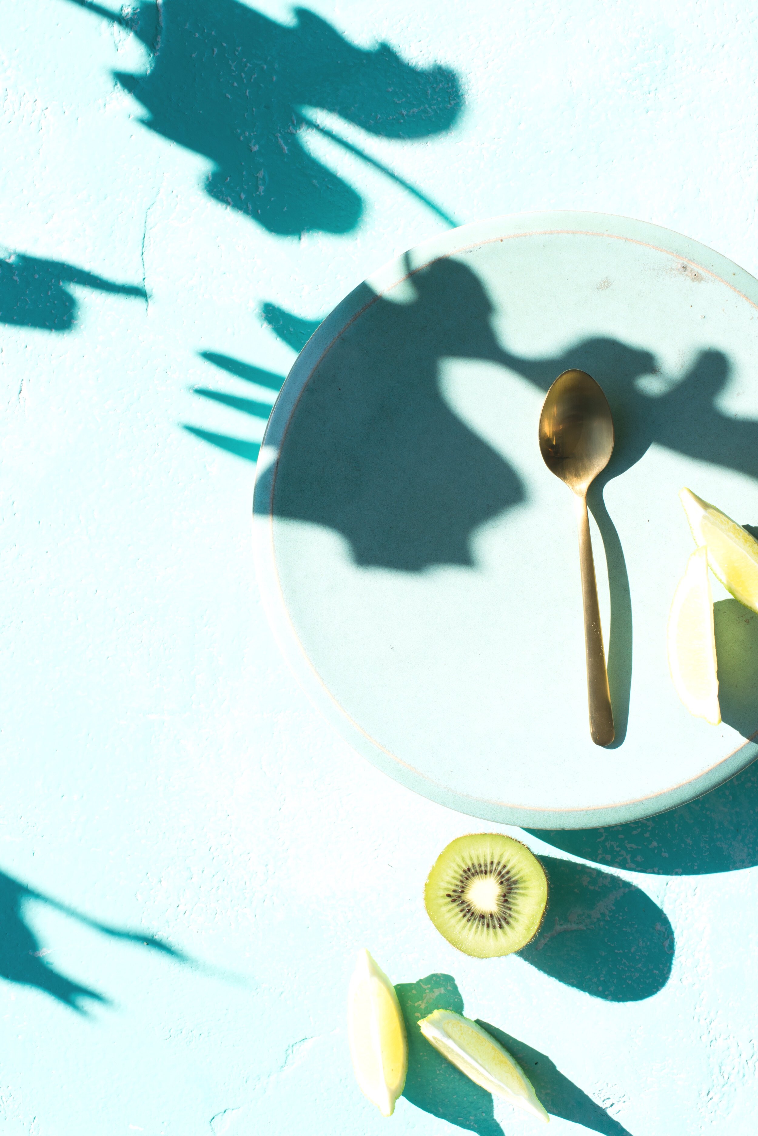 A green platter with a kiwifruit half, gold spoon, lime wedges. Natural light casts shadows of large leaves over the shot.