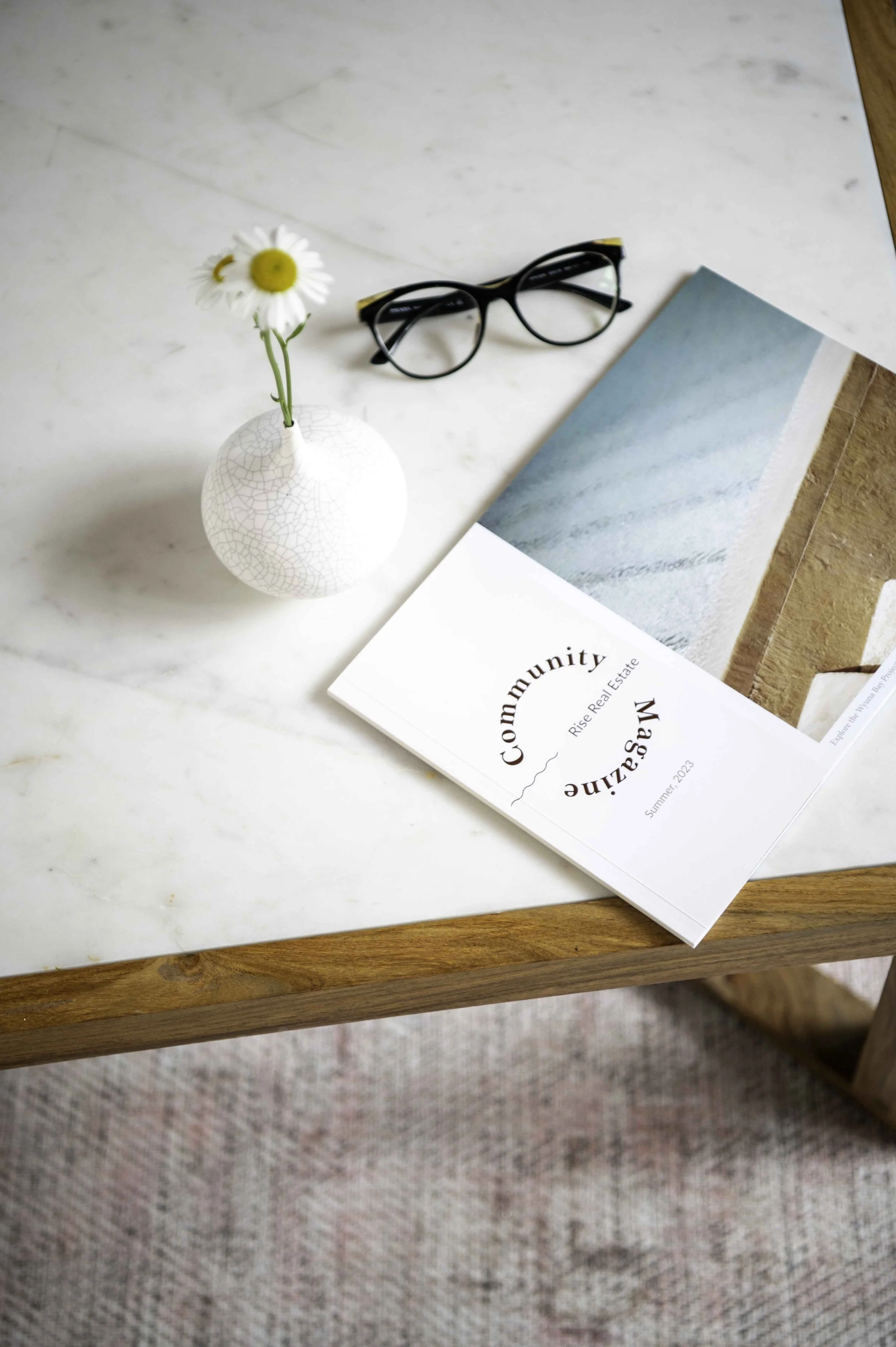 Rise Community Magazine Summer 2023 sits on a marble-top coffee table alongside a white vase with daisies and a pair of reading glasses.