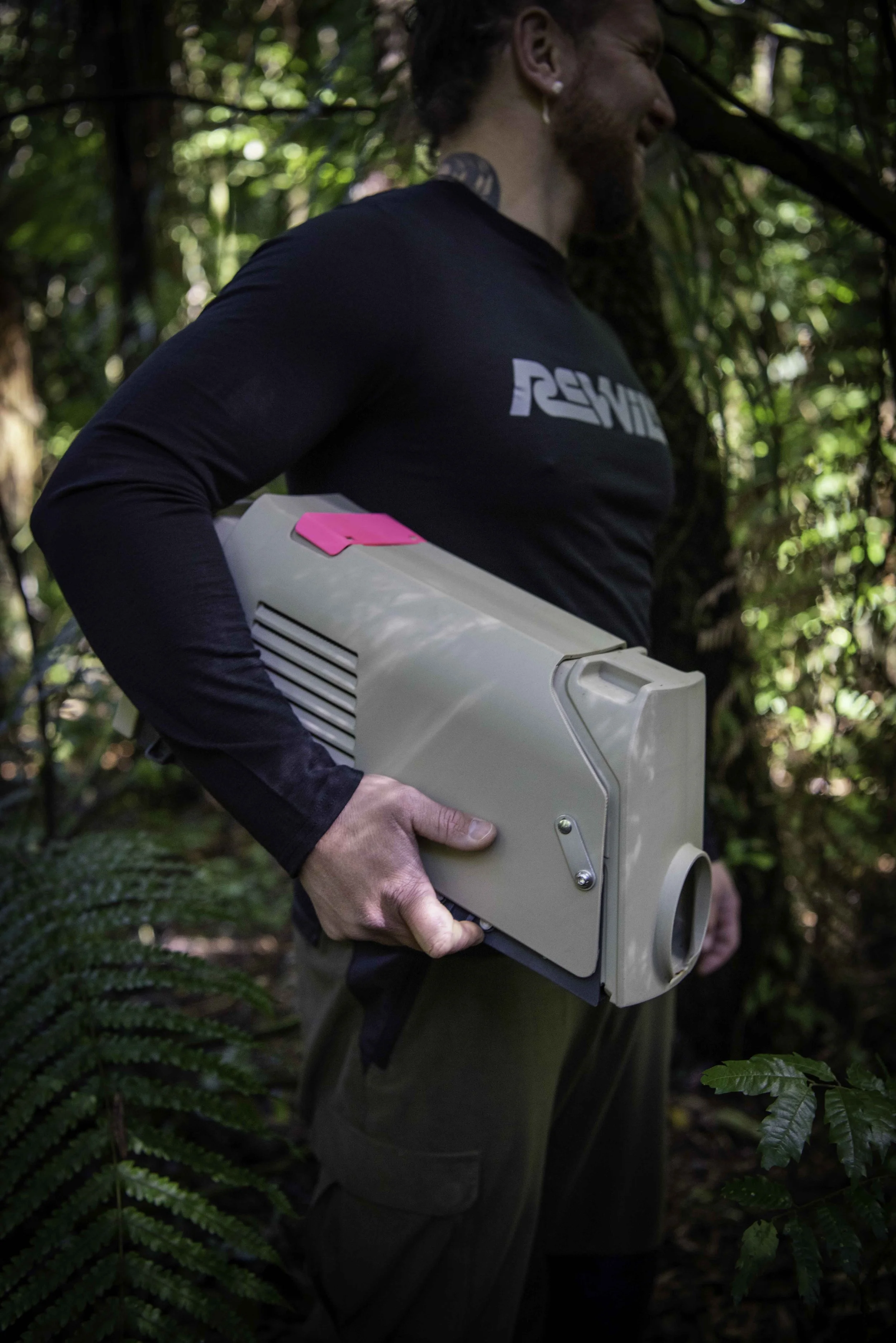 A man in a black long sleeved t-shirt holds a predator trap in the New Zealand Bush.