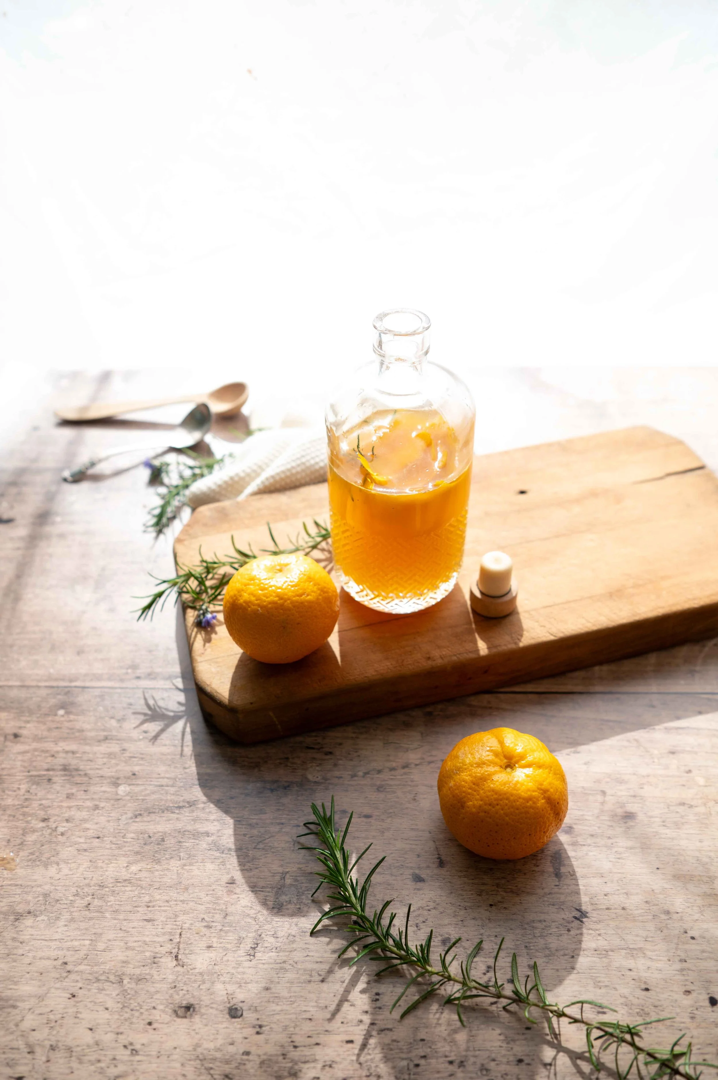 Orange & Rosemary Syrup in a glass bottle. Back-lit with oranges and rosemary sprigs in the foreground.