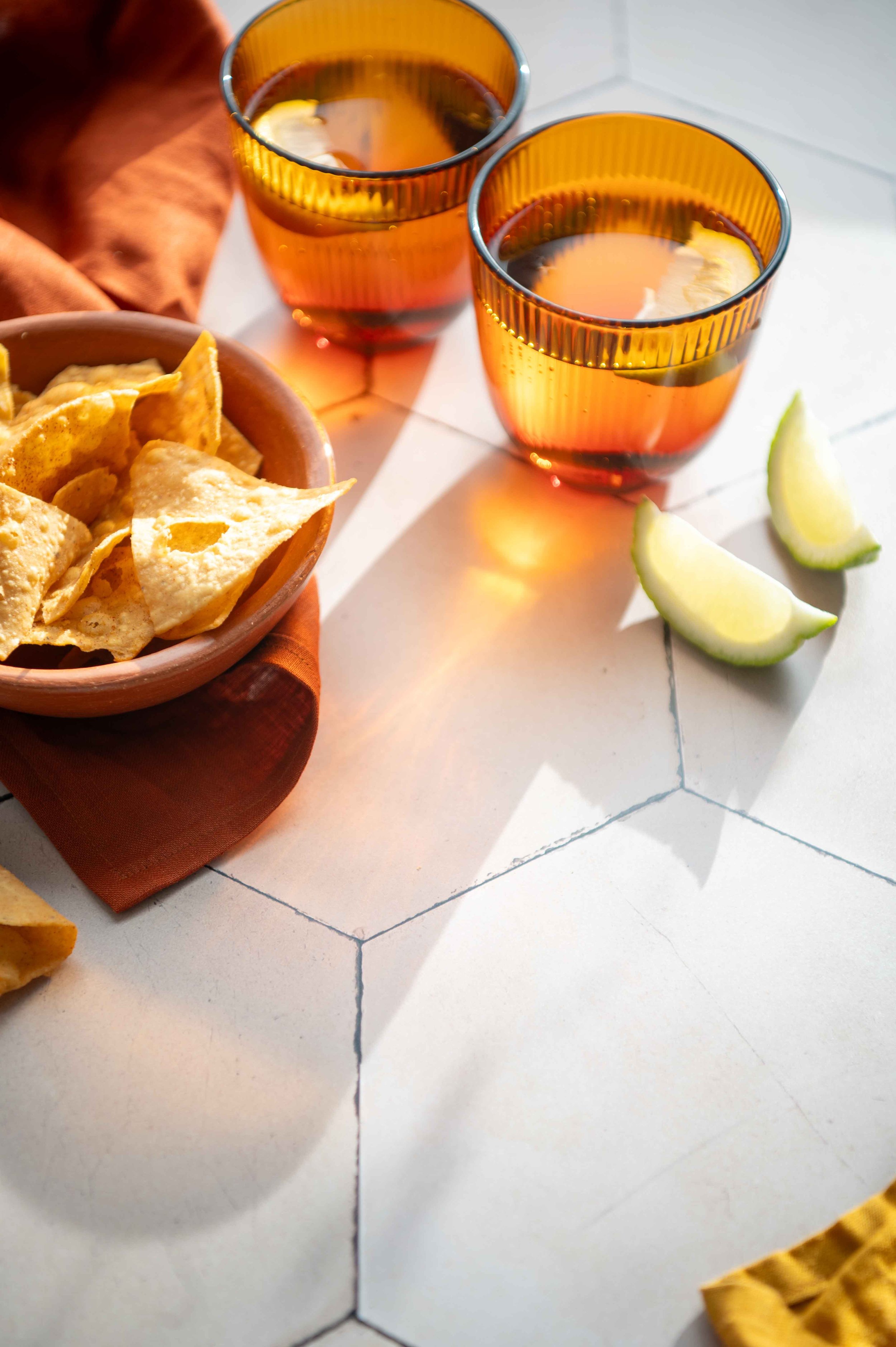 The light shines through two amber water glasses. Lime wedges, a bowl of corn chips and a rust coloured napkin.