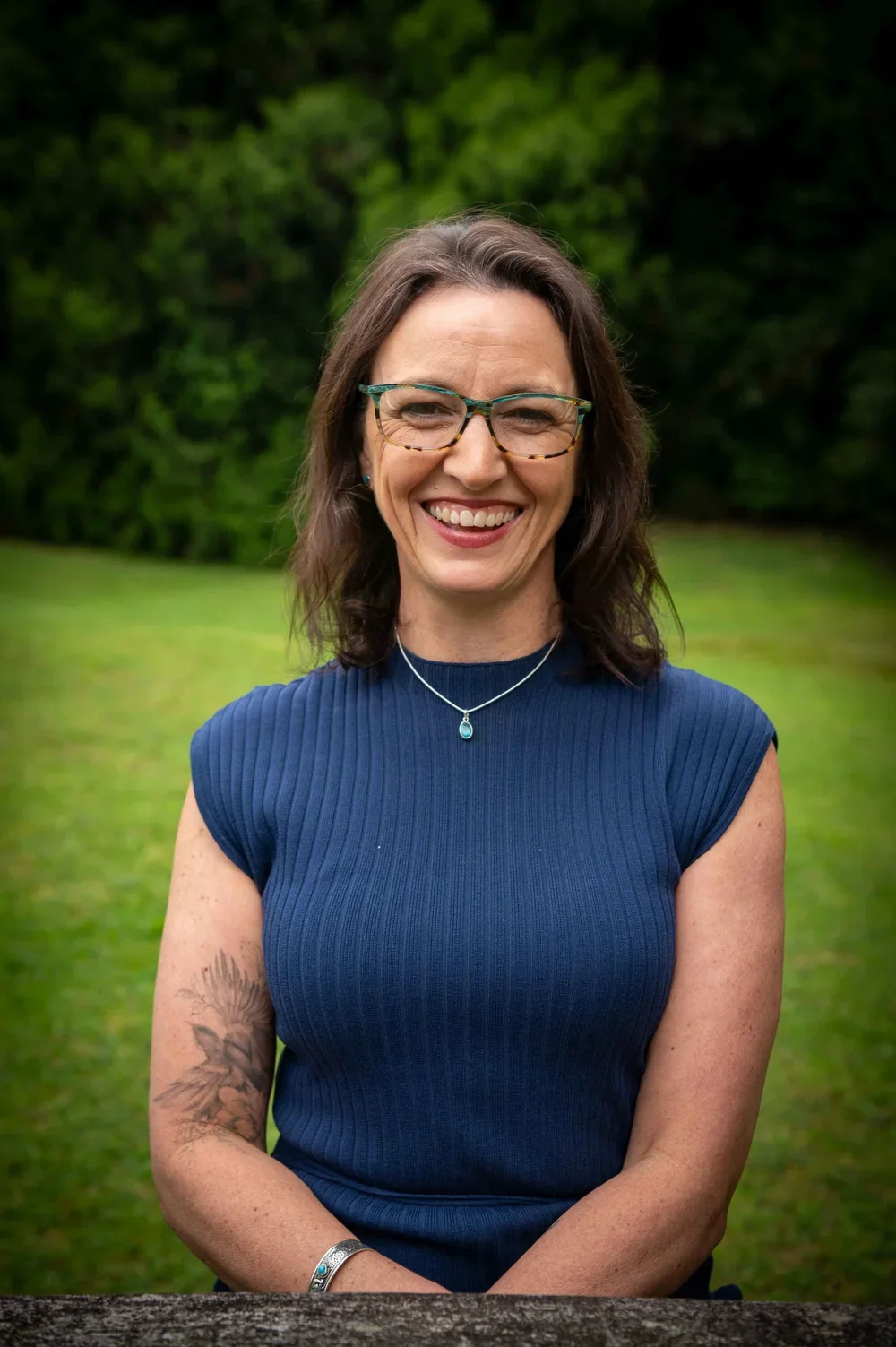 A woman with shoulder-length brown hair, glasses, and a big smile, wearing a blue sleeveless top, a necklace with a pendant, a bracelet, and a tattoo of flowers on her left arm, standing outdoors in a grassy area with green trees in the background.
