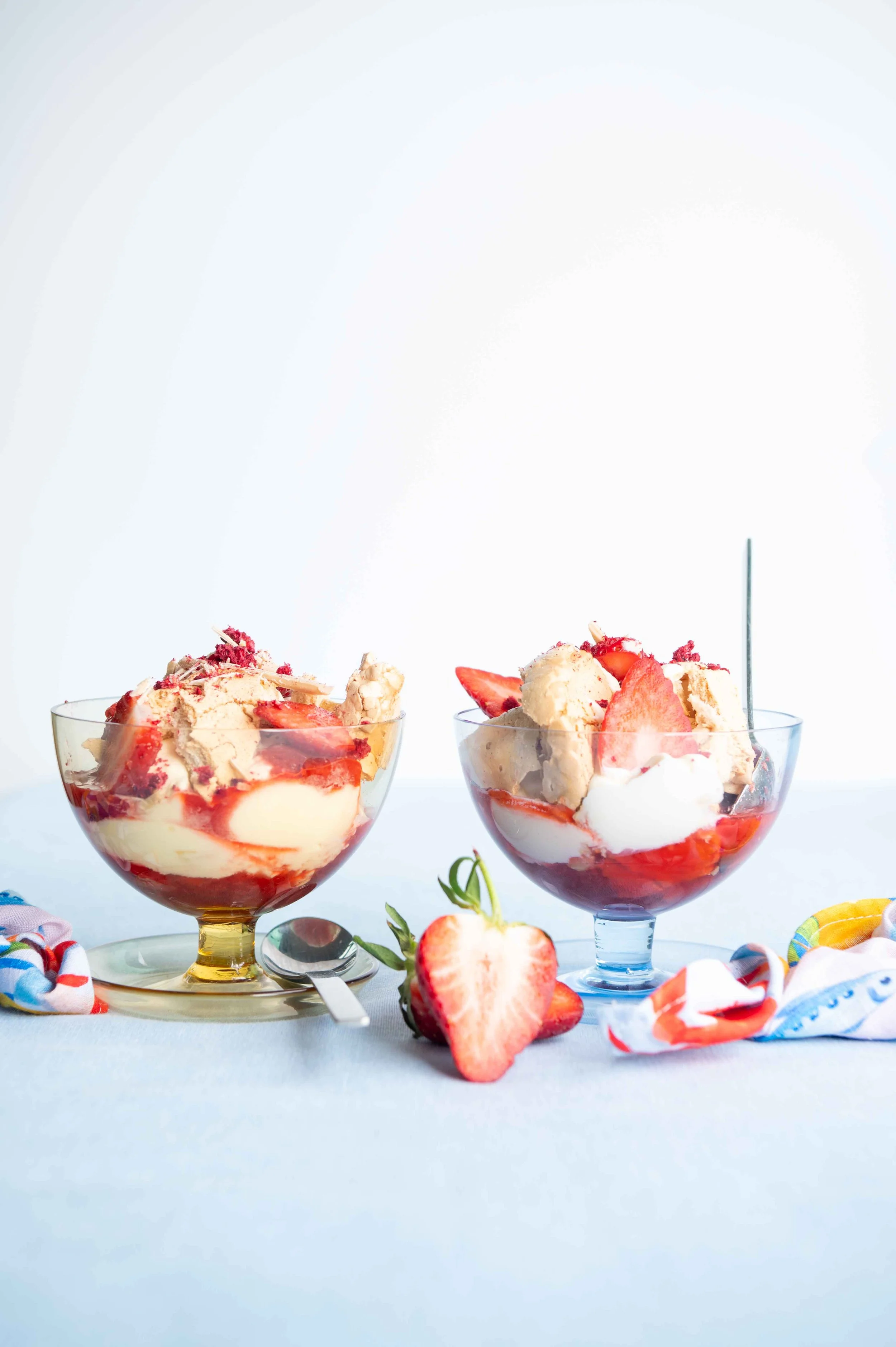 Two bowls of strawberry Eton Mess shot for Life & Leisure Magazine, set on a light blue cloth with a halved strawberry and colorful cloths around.