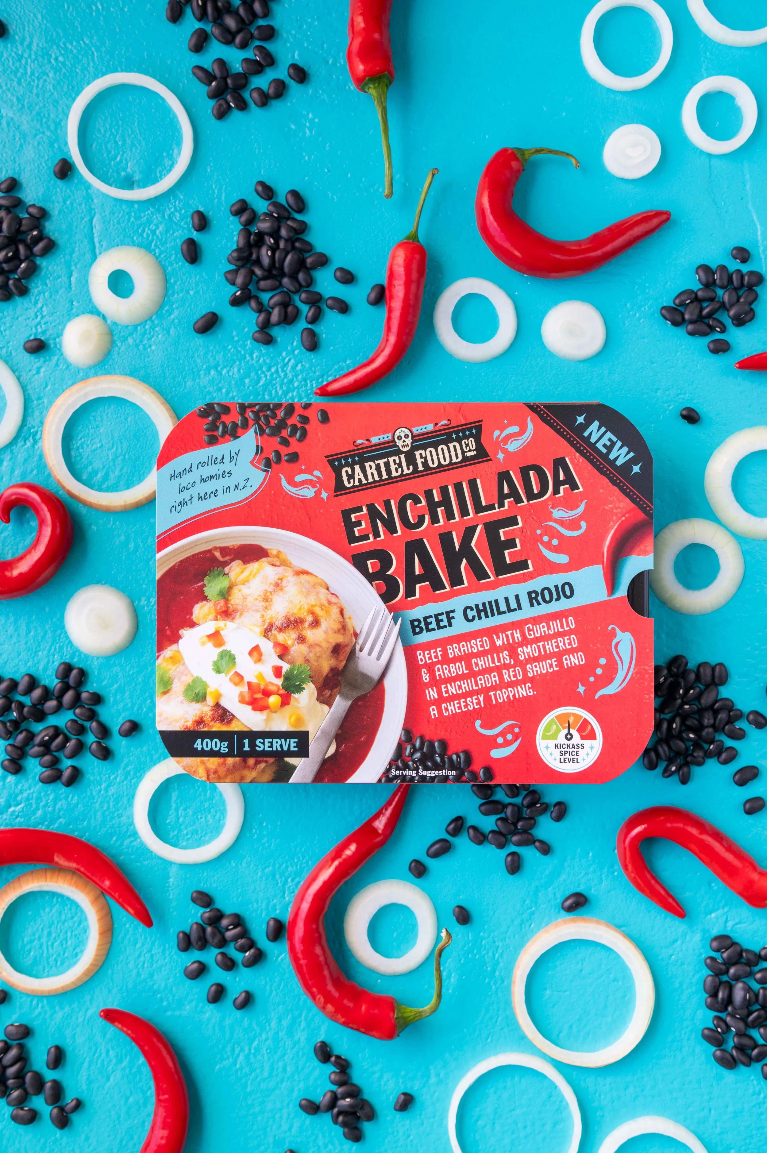 A package of Cartel Food Co. Enchilada Bake. Beef Chili Rojo flavour. Shot on a bright blue surface surrounded by red chili peppers, onion rings, and black beans.