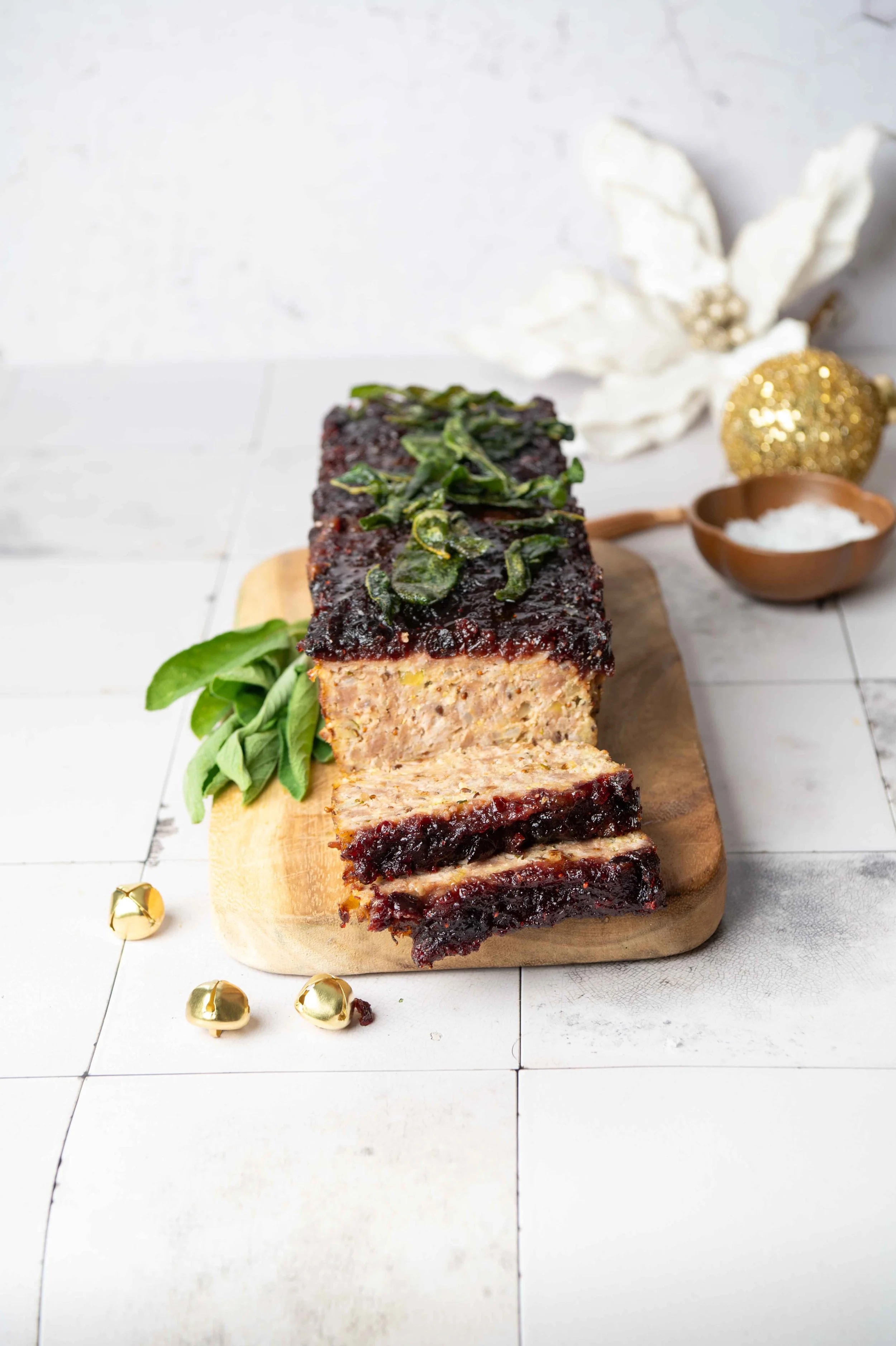 A meatloaf with a dark glaze and green herbs on top, placed on a wooden cutting board, with some fresh green herbs beside it, festive decorations in the background, including a poinsettia flower and gold ornaments.