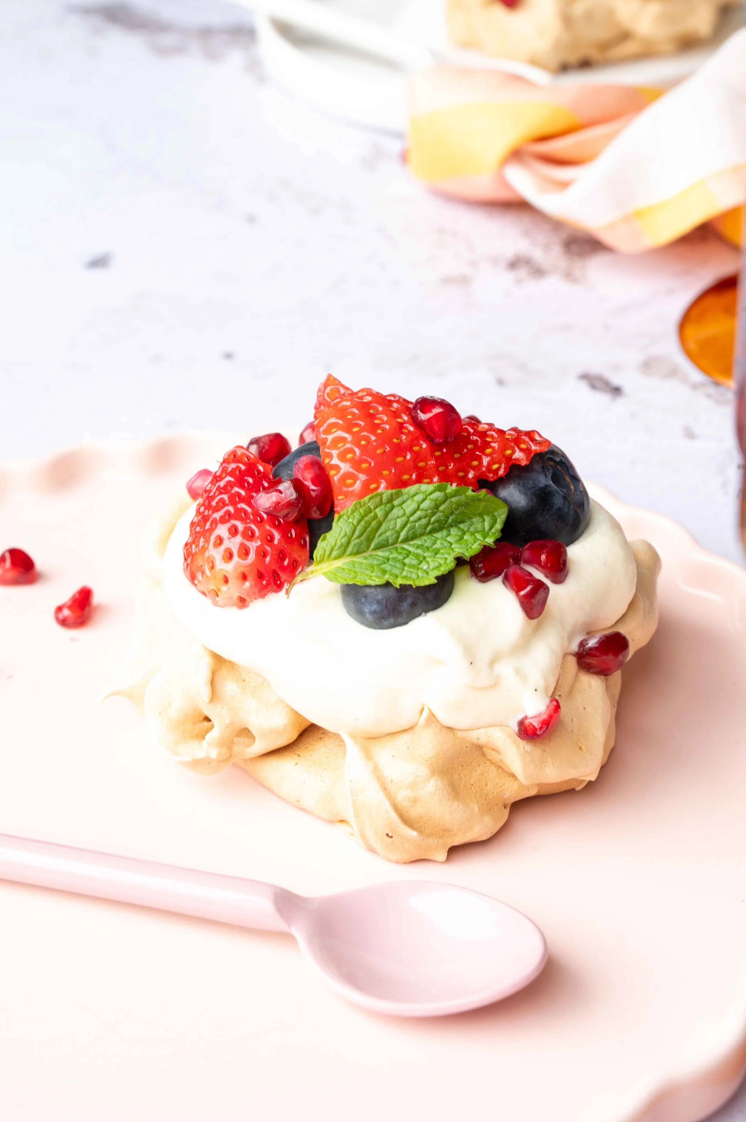 A dessert with meringue topped with whipped cream, strawberries, blueberries, pomegranate seeds, and a mint leaf on a pink plate.