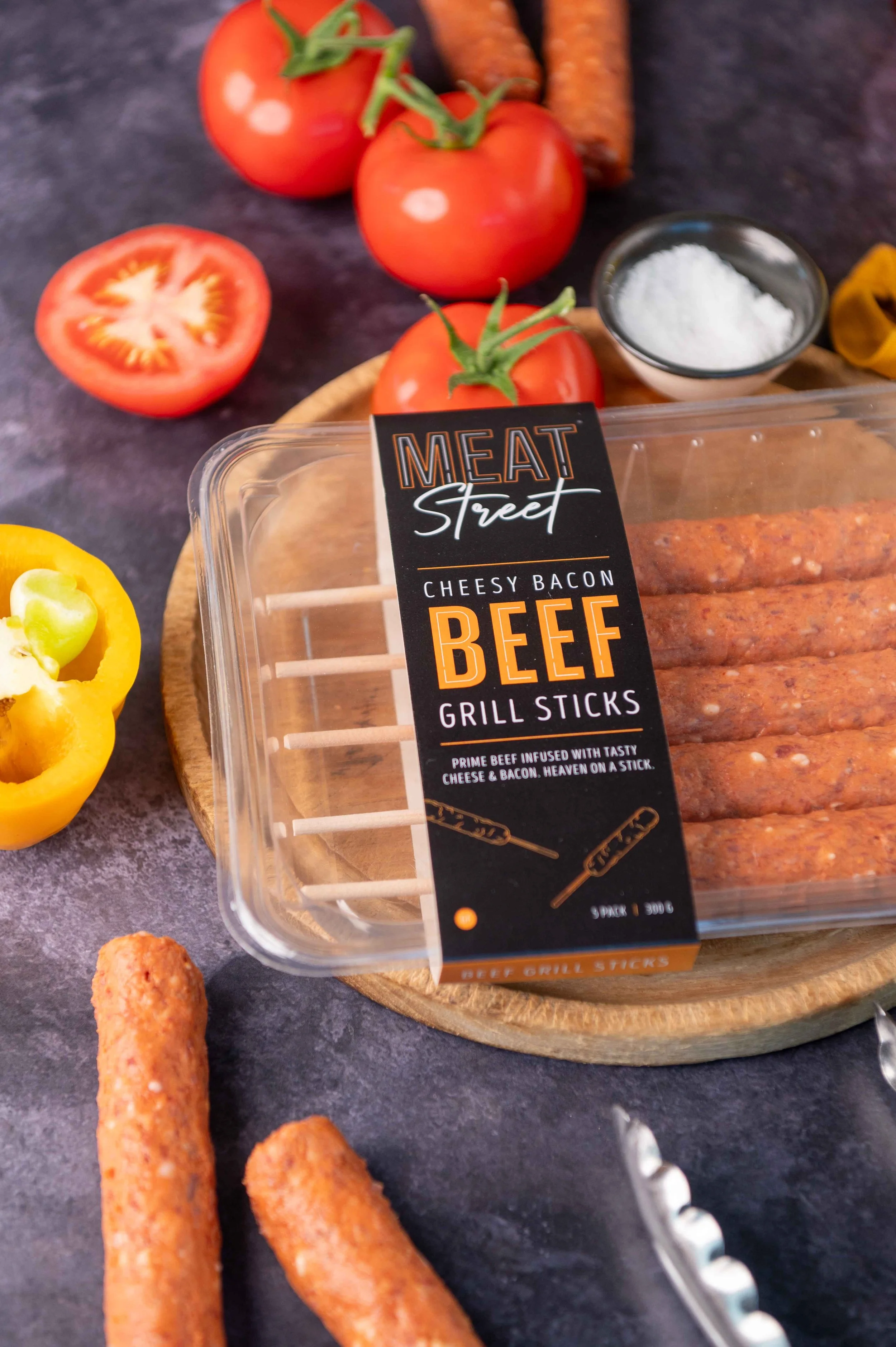 Package of Meat Street bacon beef grill sticks surrounded by tomatoes, a yellow bell pepper, some salt, and sausages on a dark surface.