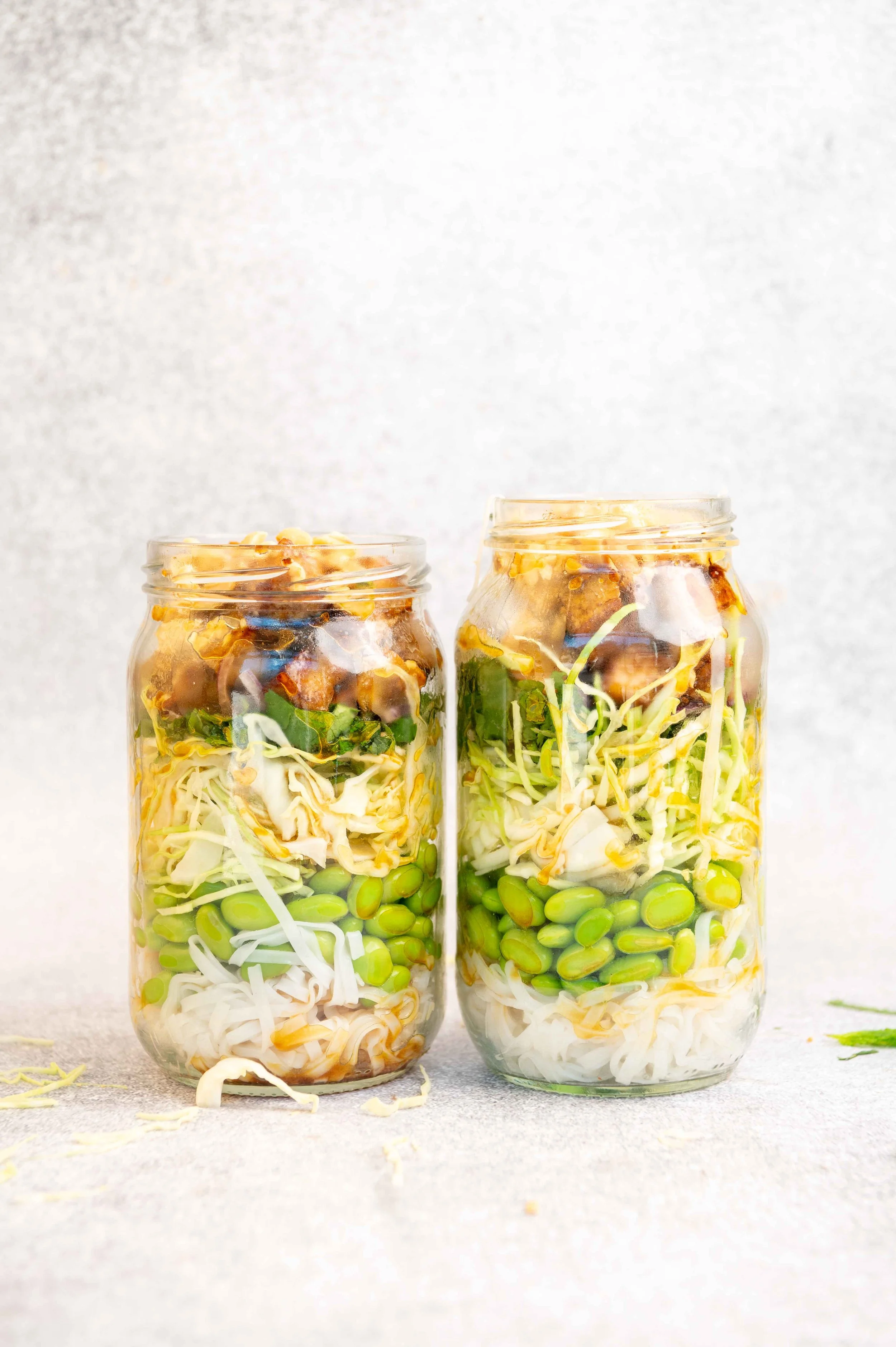Two Mason jars filled with layered ingredients including shredded cabbage, green beans, and cooked meat, ready for pickling. Tofu Noodle Salad for Life & Leisure Magazine.
