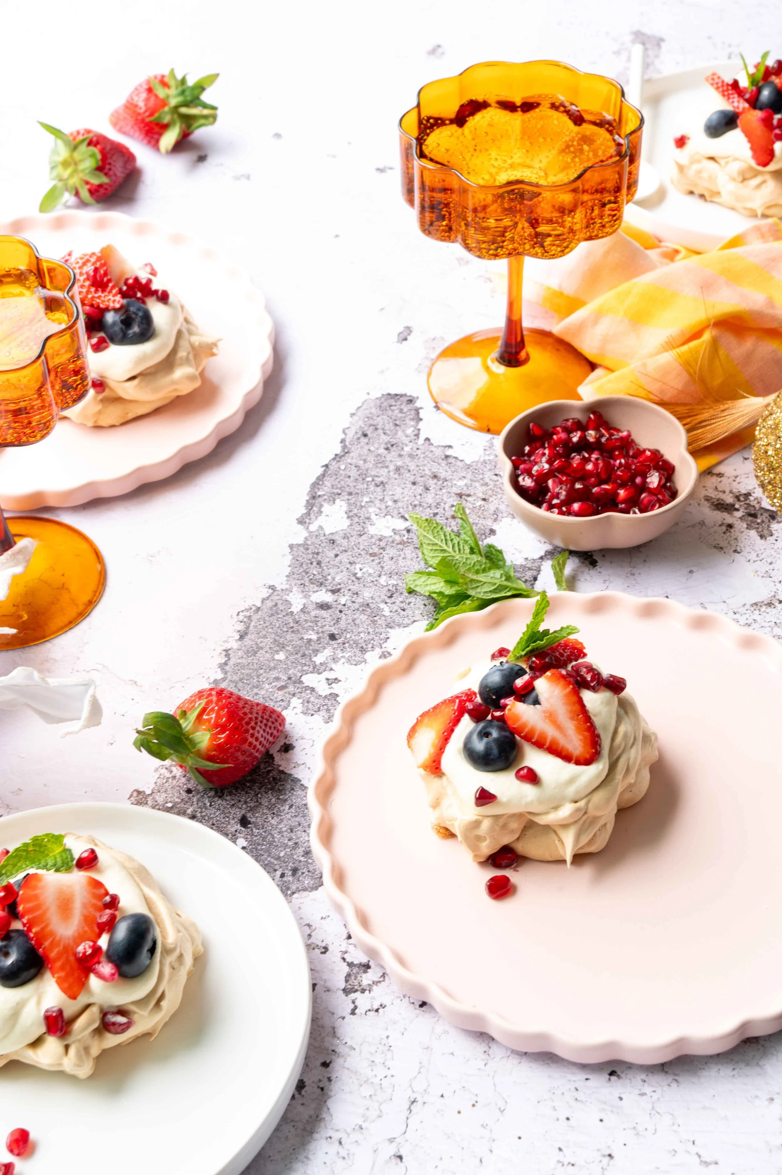 Brown Sugar Meringues are arranged on three plates and are decorated with cream, strawberry slices, blueberries and pomegranate arils. There are two amber champagne glasses, napkins and strawberries and mint leaves on the white textured background.
