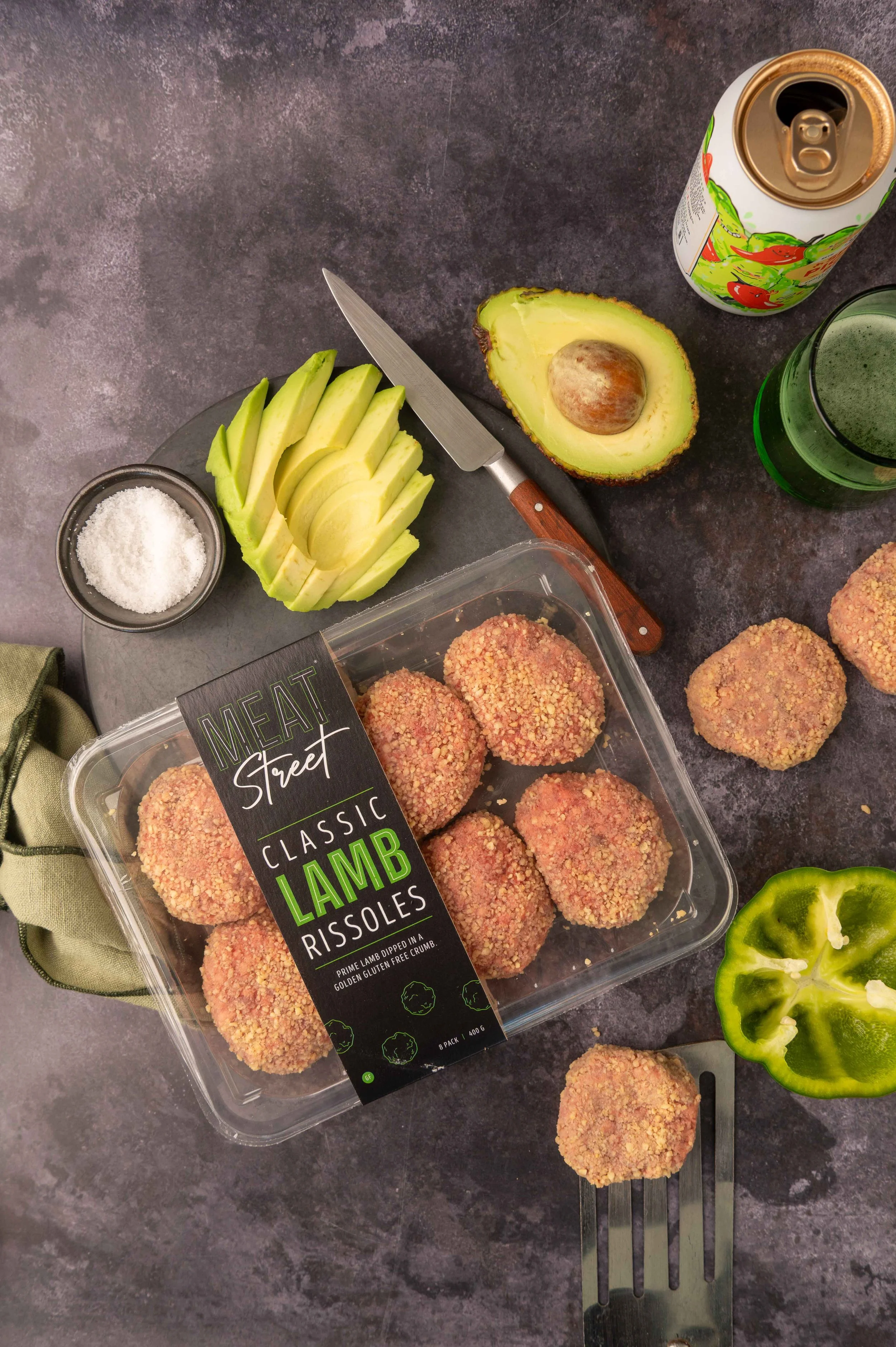 Packaged Meat Street lamb rissoles with sliced avocado, half a bell pepper, salt, beer in a glass with the can. Shot on a dark grey background.