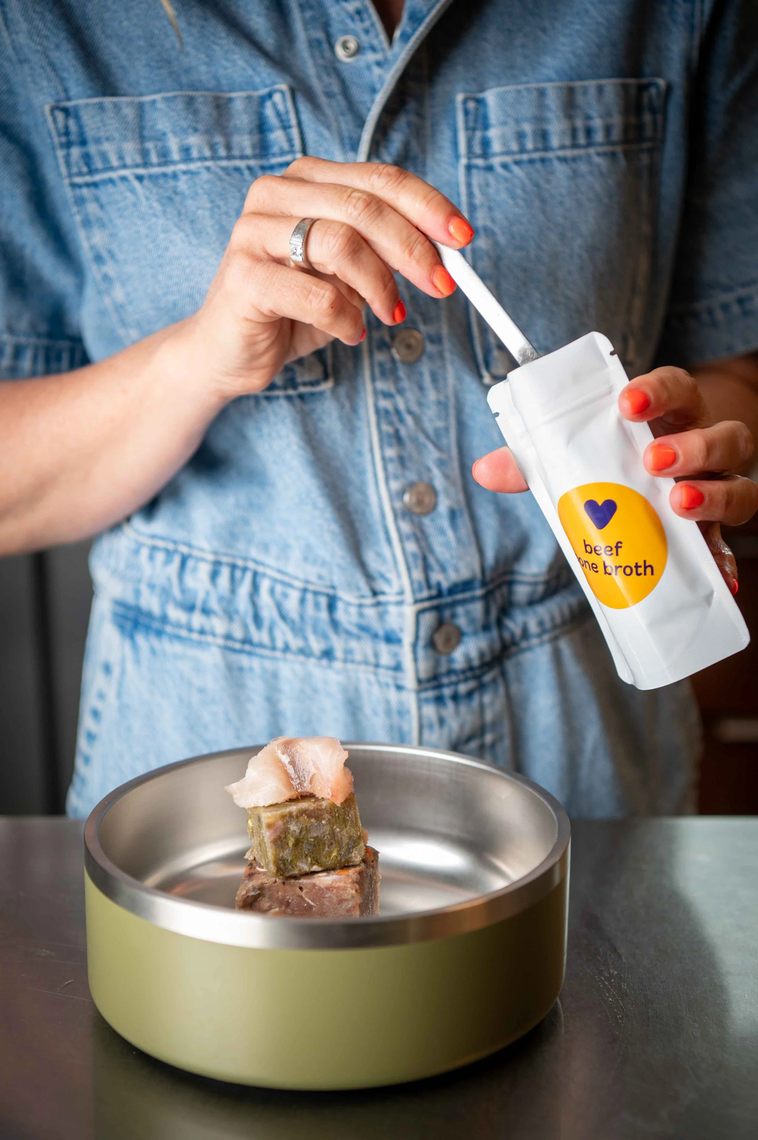 A woman in a denim jumpsuit with orange nails sprinkles bone broth powder over a serving of My Raw Pet food.
