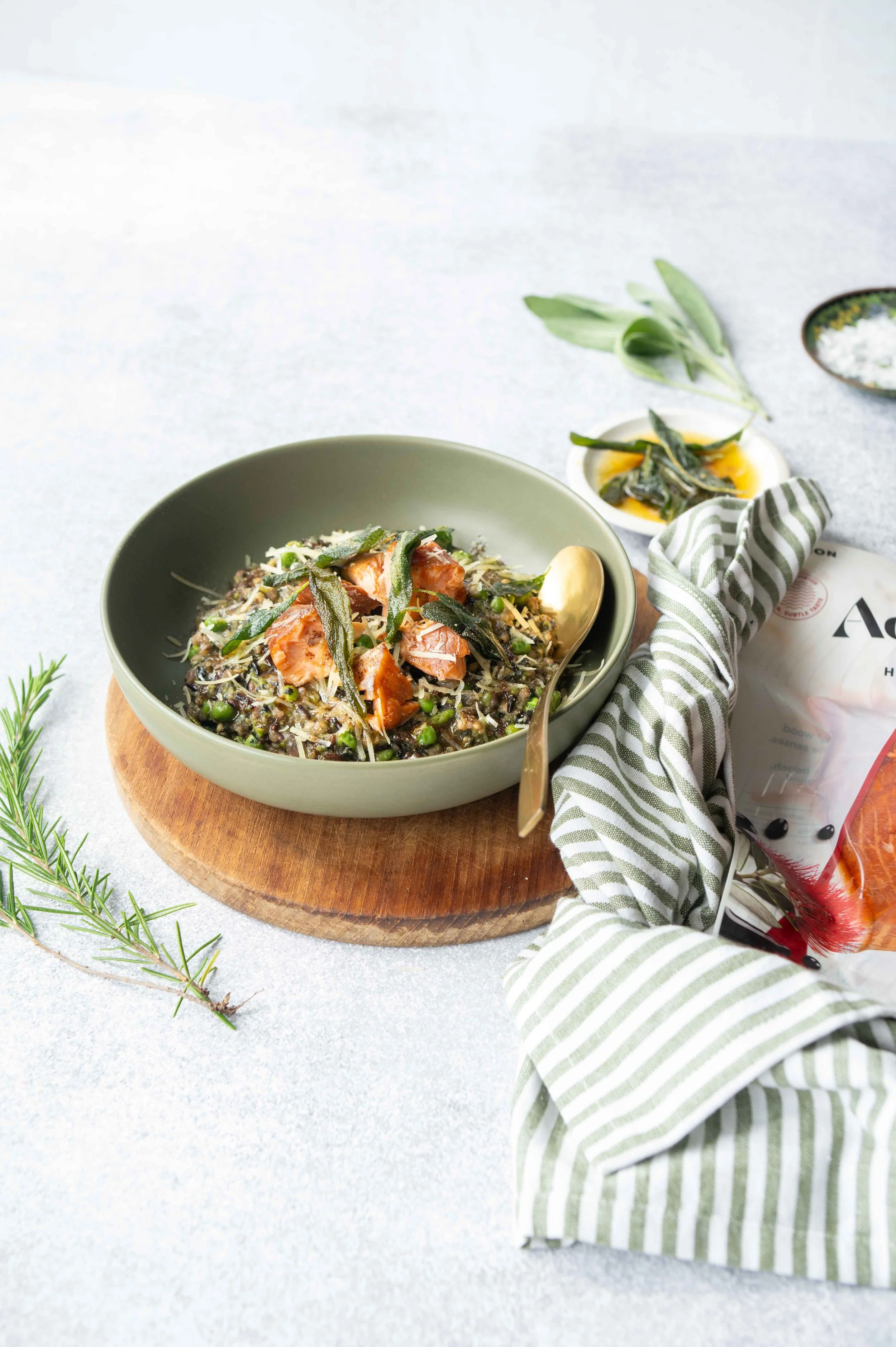 Aoraki Smoked Salmon & Kale Risotto. Shot on a light grey background in a sage green bowl. A packet of Aoraki Salmon coming into shot. Sprigs or rosemary and a green and white striped napkin.