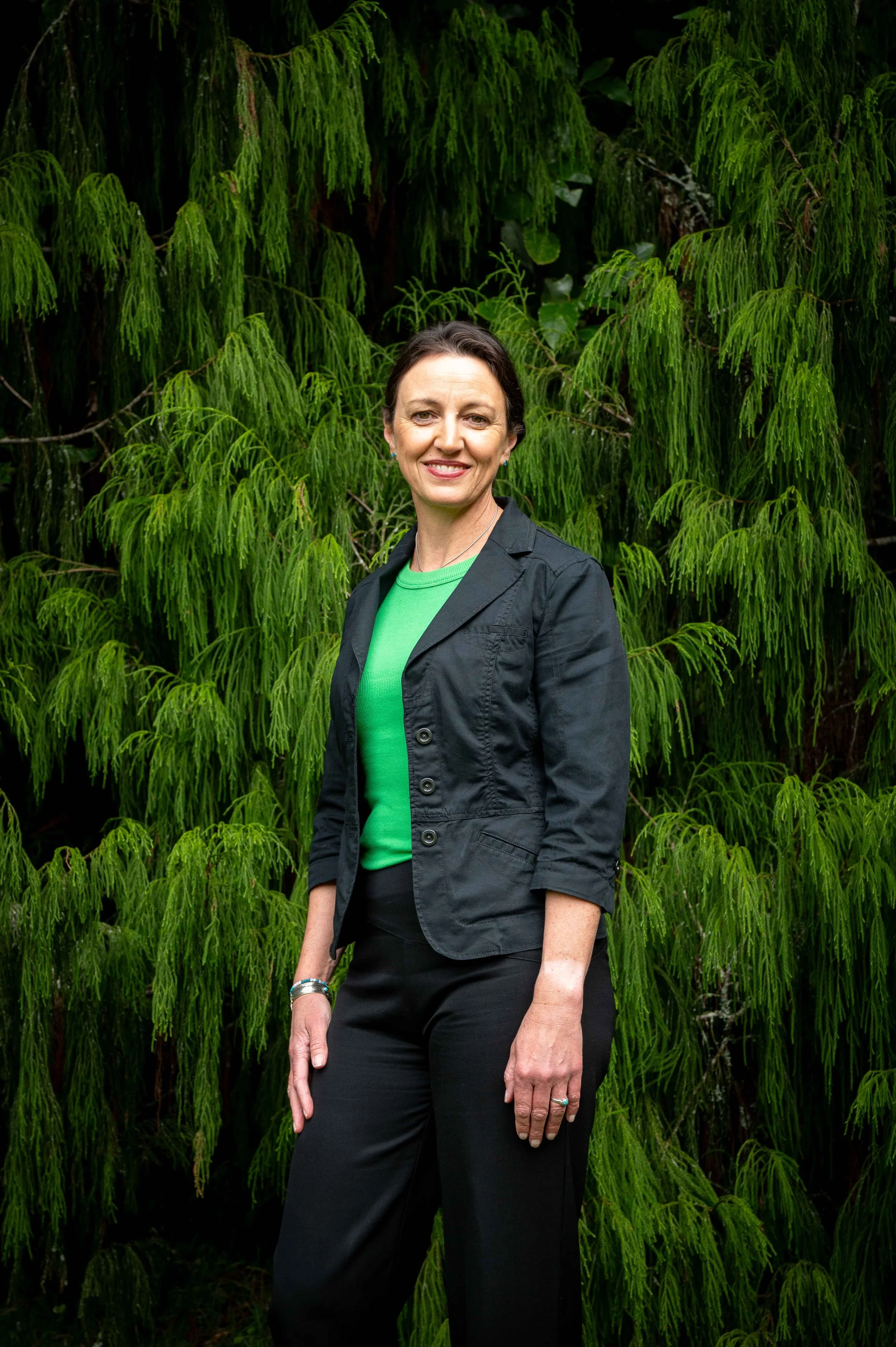 A woman standing outdoors in front of lush green trees, wearing a black jacket and black pants, smiling at the camera.