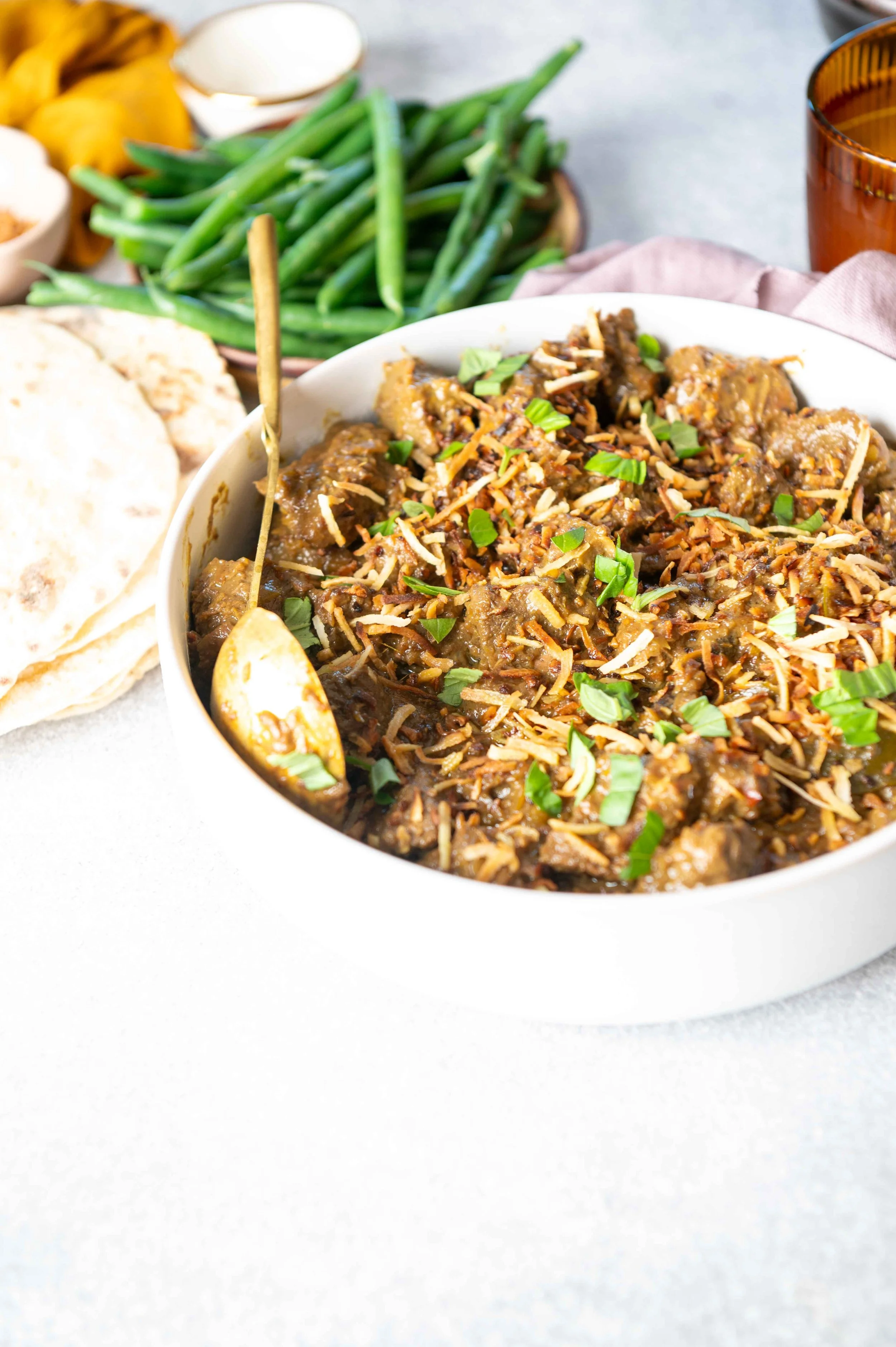 A bowl of beef chili topped with shredded cheese and chopped green onions, with green beans, flatbread, and a cup in the background.