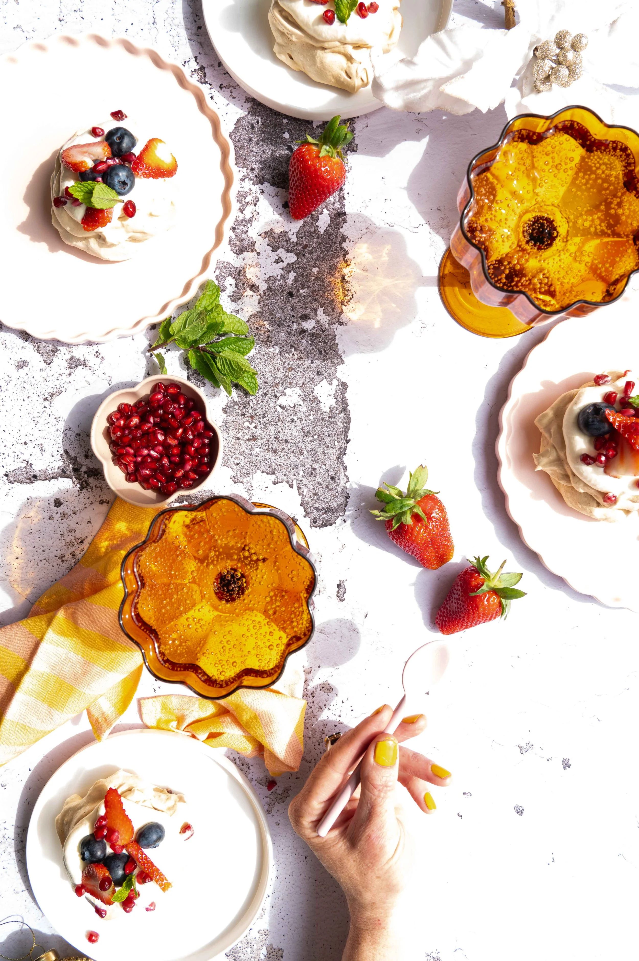 Table set with desserts, strawberries, pomegranate seeds, blueberries, glass of sparkling beverage, hand with yellow nail polish holding a spoon, and scattered strawberries on a white surface.