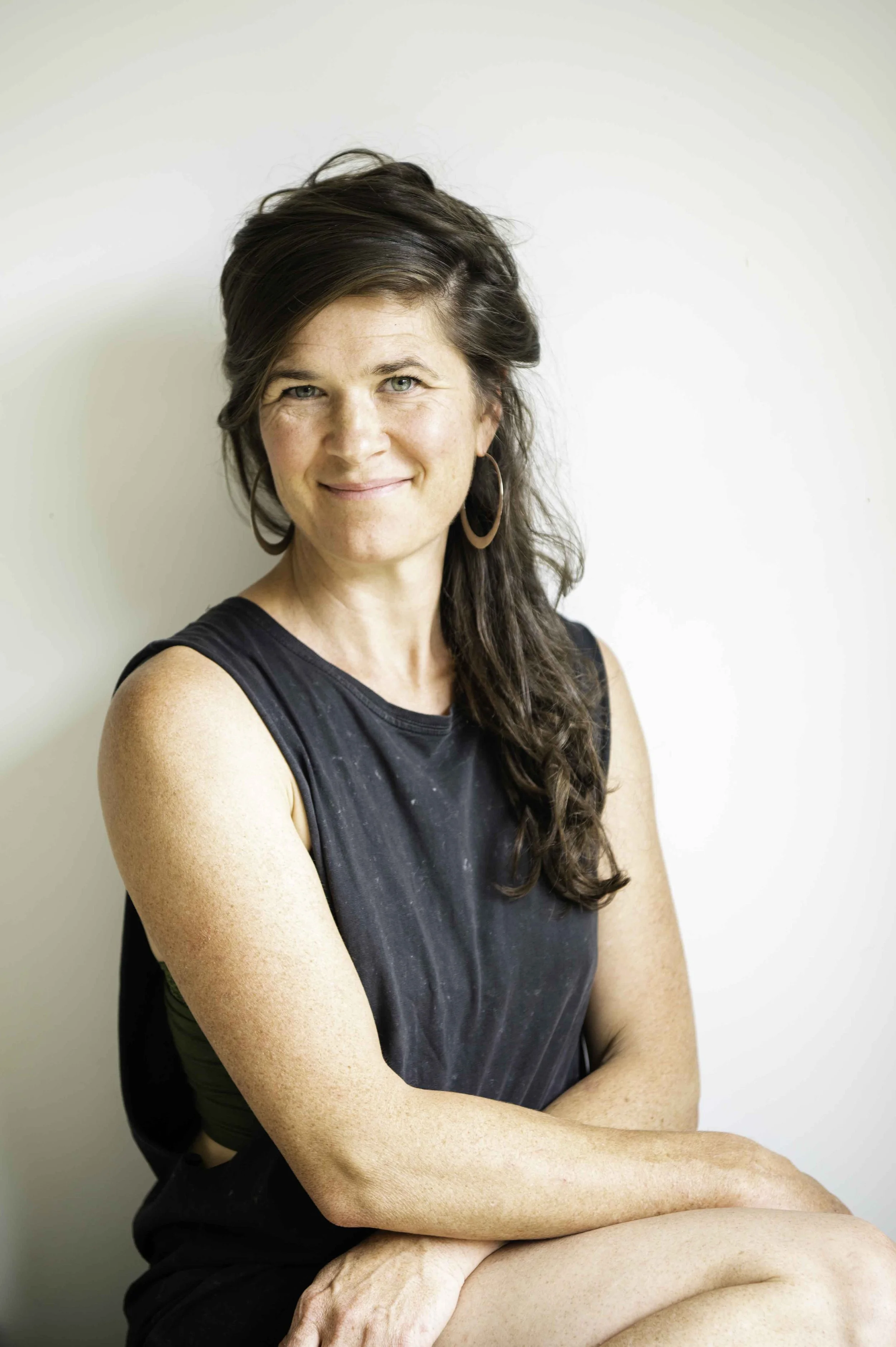Picture of a woman shot against a neutral background. Long dark brown hair falls over her left shoulder. She wears a grey singlet and large silver hoop earring.