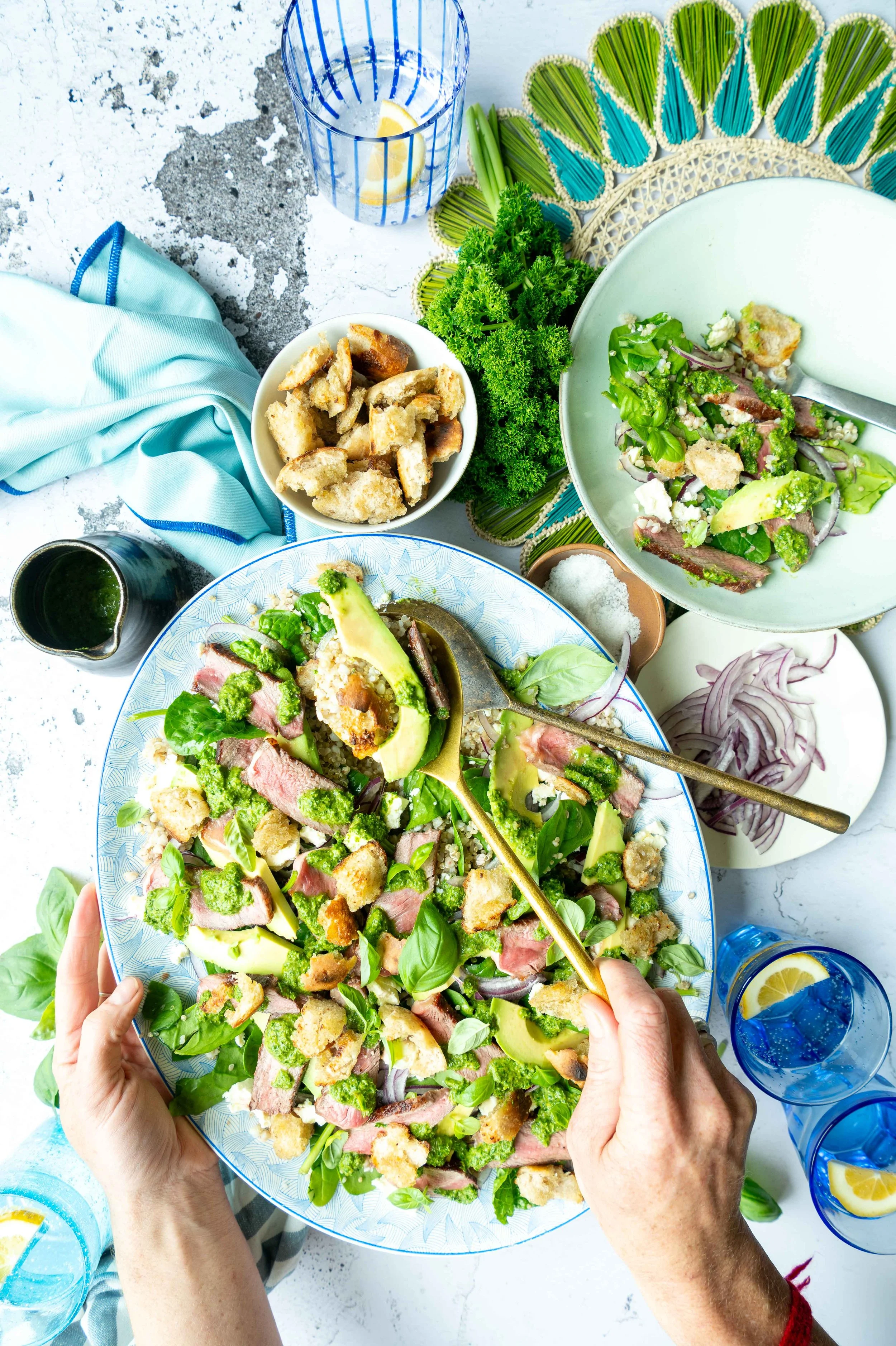 Overhead view of a fresh green salad with sliced steak, avocado, and croutons on a large decorative plate, surrounded by bowls of croutons, sliced onions, and salad ingredients, with two glasses of water with lemon slices.
