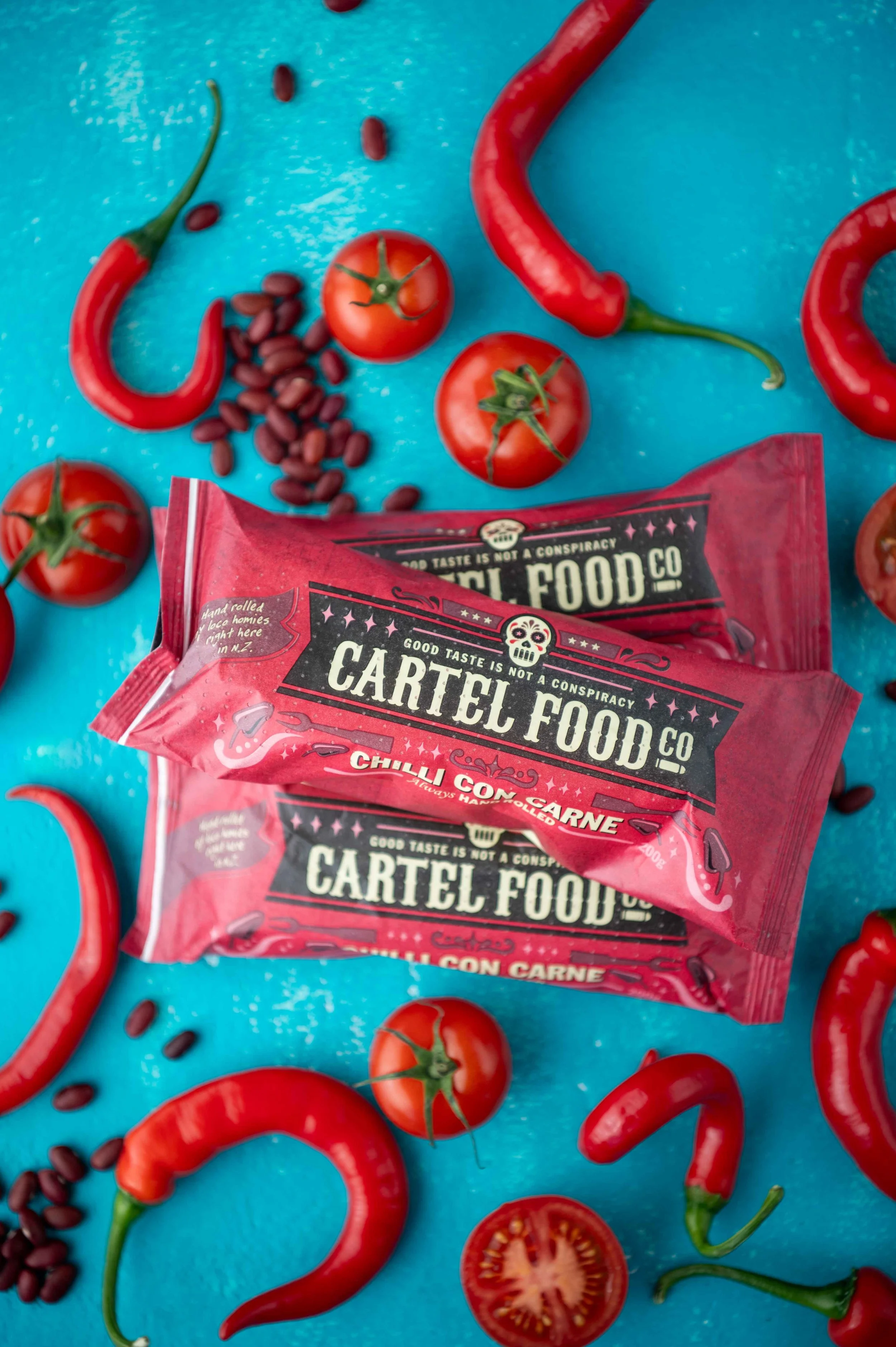 Three packets of Cartel Food Co beef burritos surrounded by red chili peppers, cherry tomatoes, and black beans.