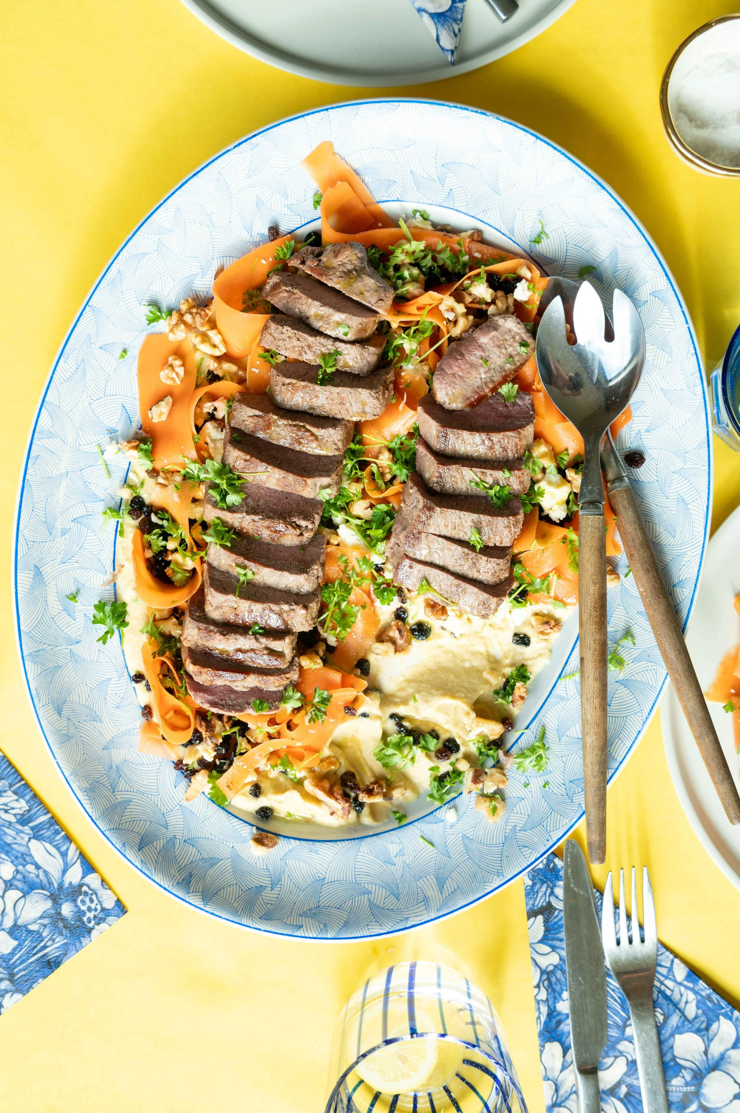 A plate with sliced cooked steak, carrot ribbons, fresh herbs, roasted vegetables, and mashed potatoes with gravy, set on a yellow table.