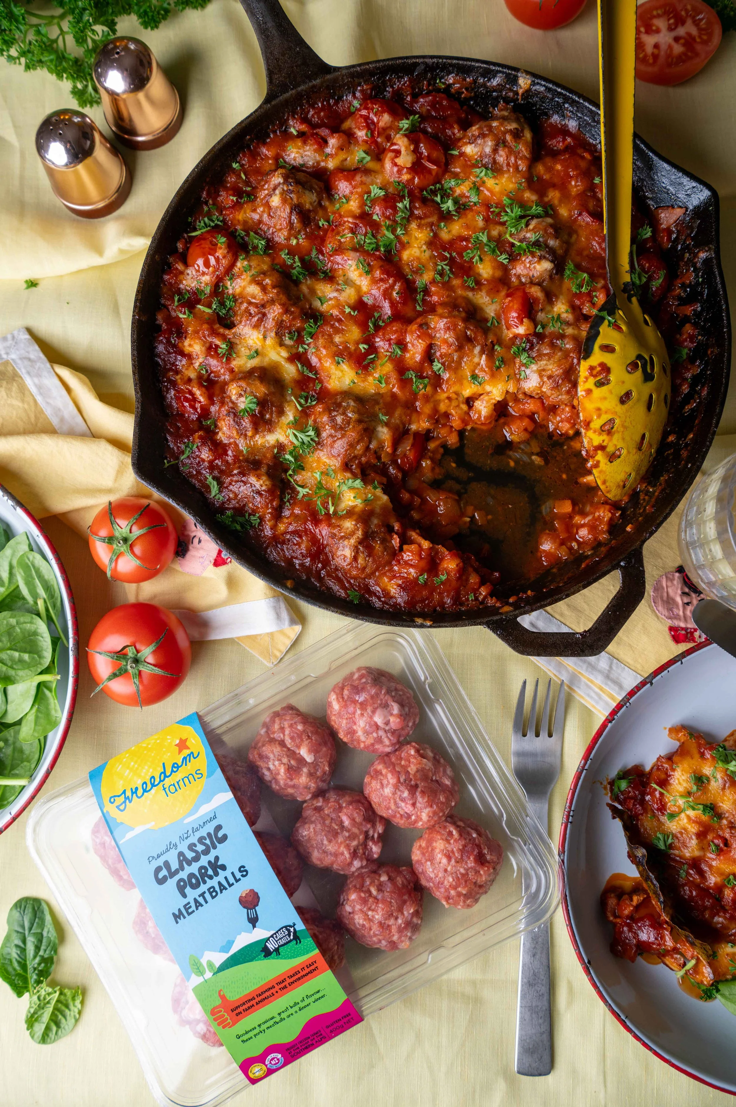 A skillet of cooked meatball casserole with tomato sauce, cheese, and herbs, surrounded by fresh tomatoes, spinach salad, and a package of pork meatballs on a table.