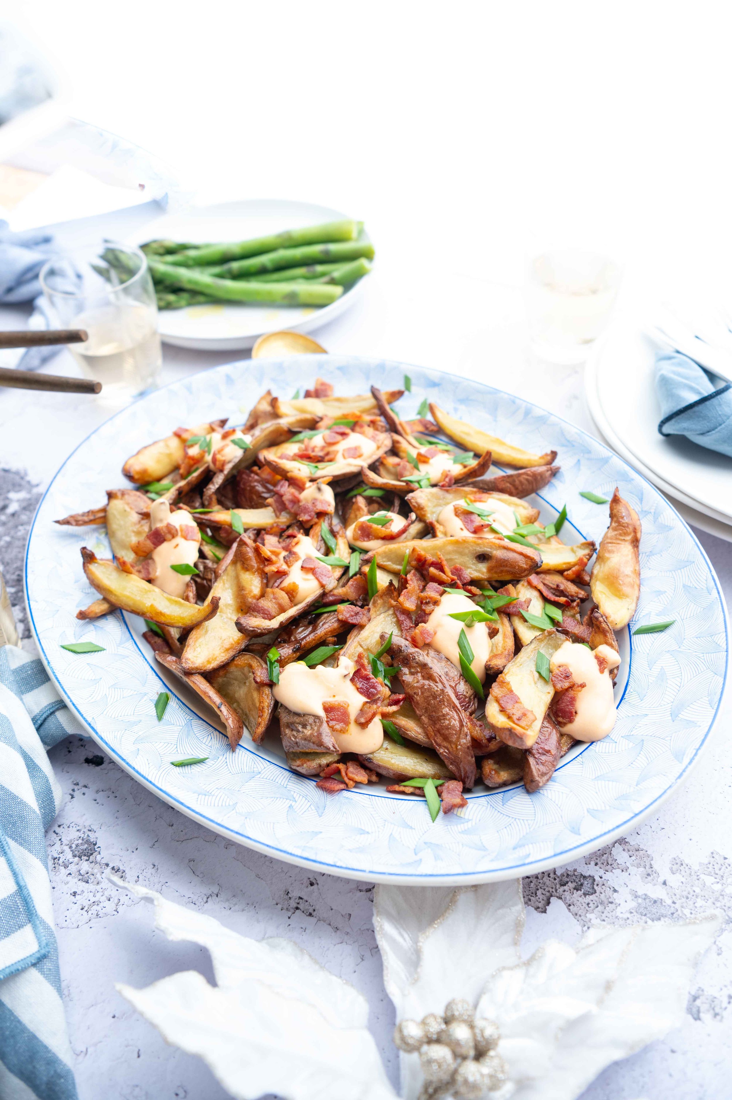 A platter of loaded potato wedges with bacon, cheese, and green onions, served on a decorative white plate on a white table, with asparagus and drinks in the background.