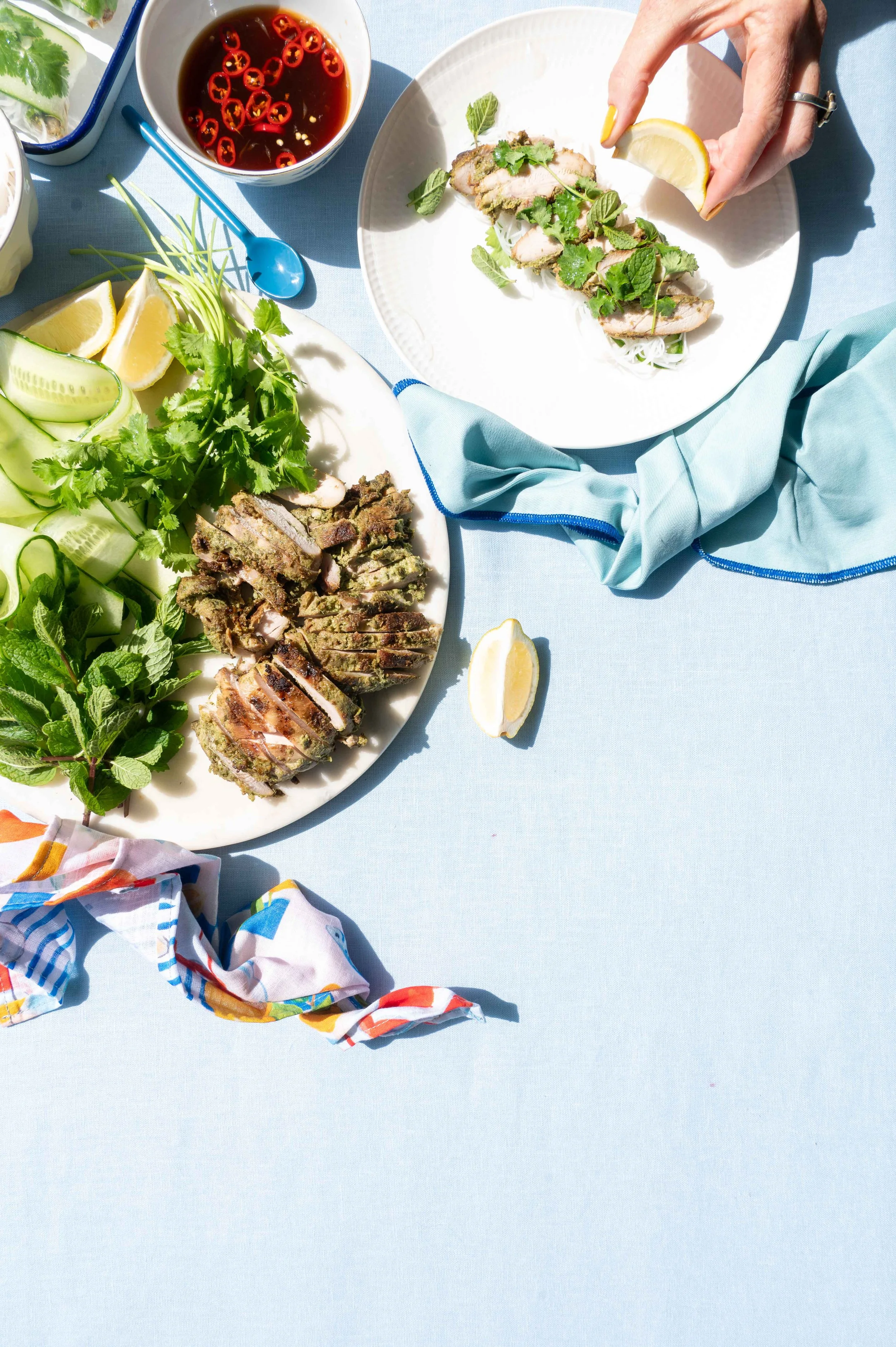 A table with Vietnamese food, including sliced grilled meat, fresh herbs, cucumber, lemon wedges, and a bowl of dipping sauce topped with sliced chili peppers. Rice paper rolls for Life & Leisure Magazine.