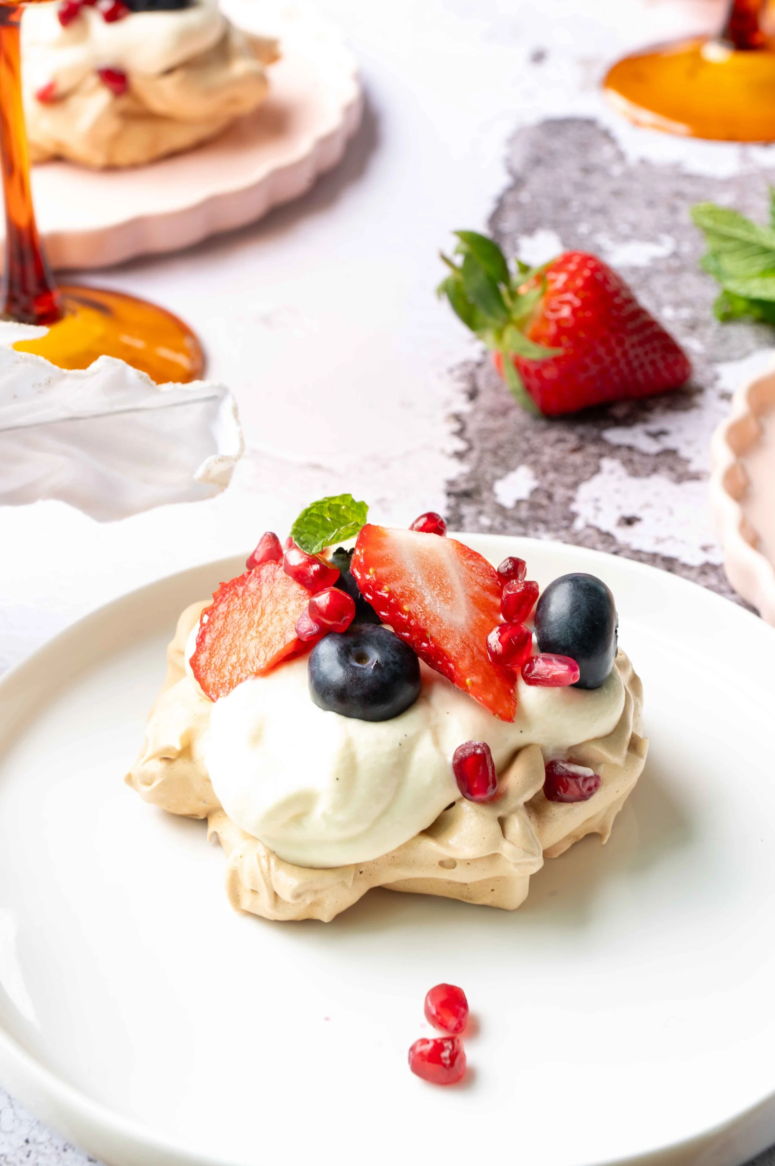 Brown sugar Vanilla Meringues sit on pink plates with strawberries on a white background. Mint sprigs and amber champagne glasses sit next to them.