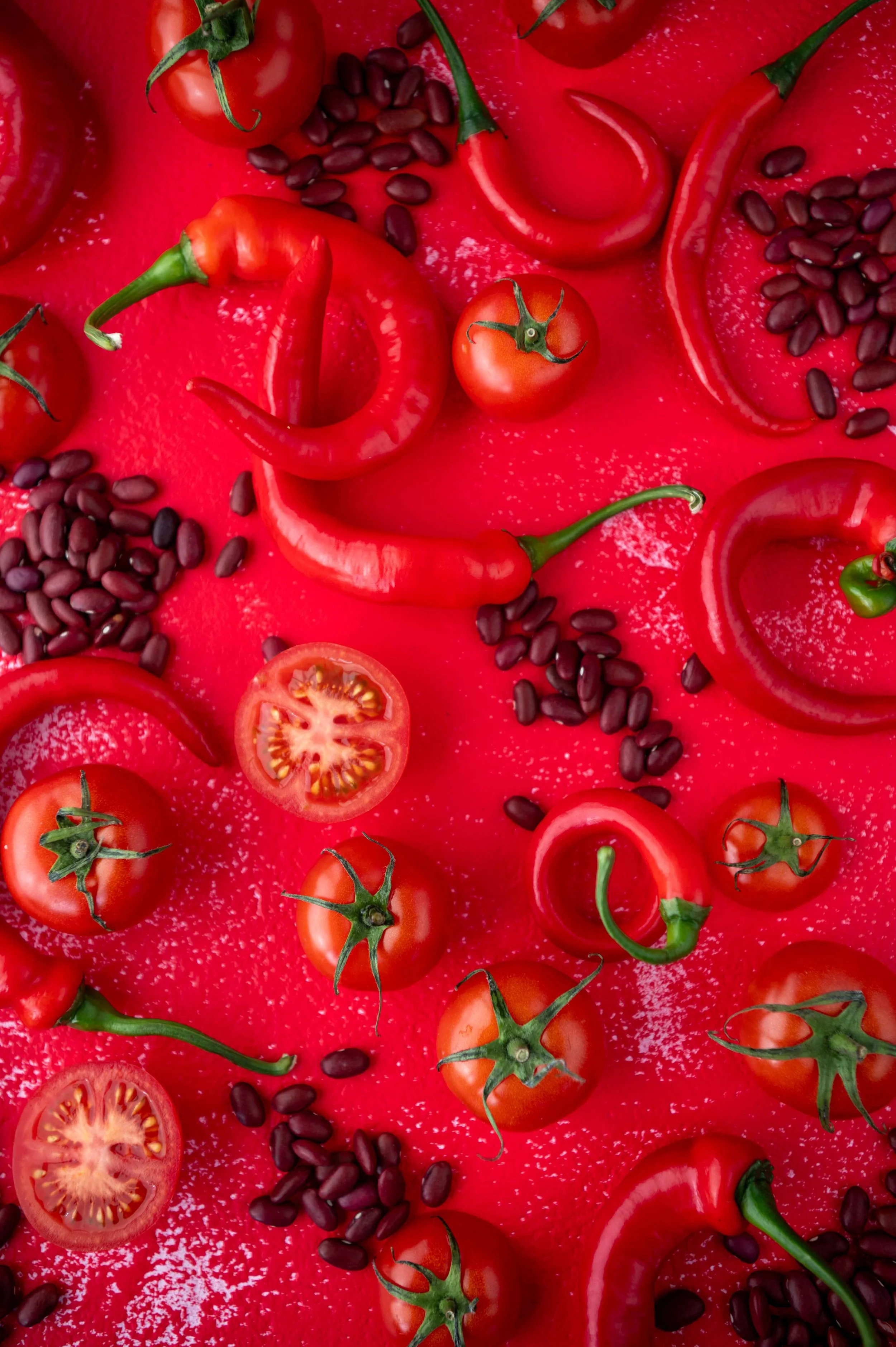 Red tomatoes, red chilis and black beans scattered on a red surface.
