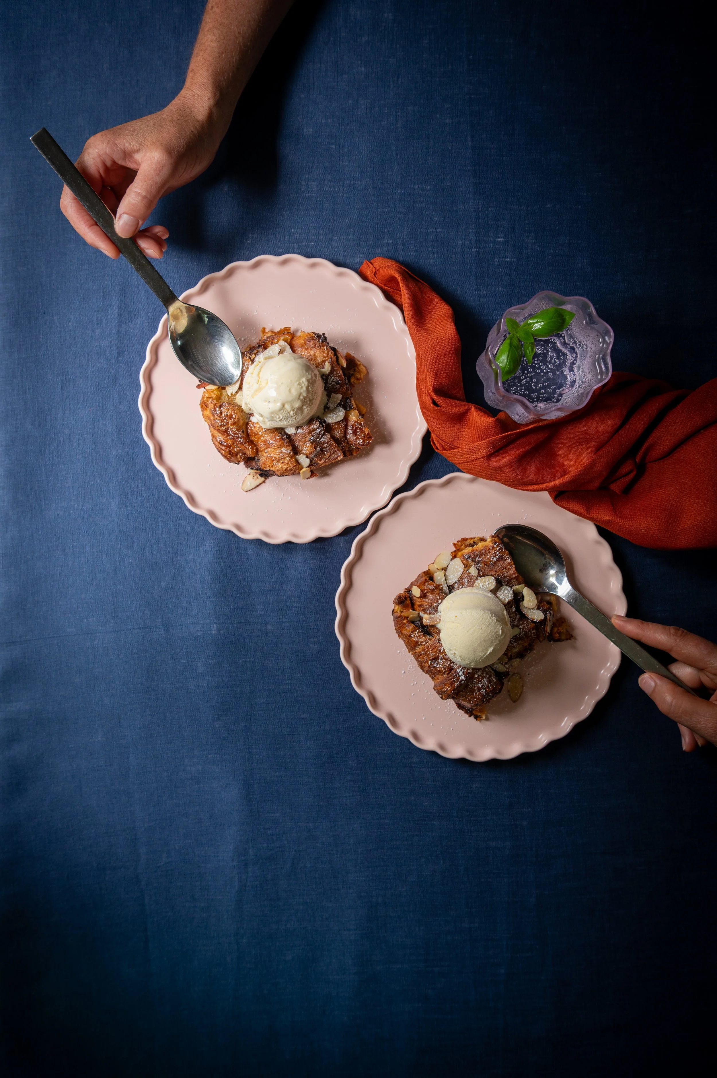 Two pink plates with croissant & chocolate pudding topped with vanilla ice cream and almond slices with a glass of water beside them, on a dark blue table with orange cloth.