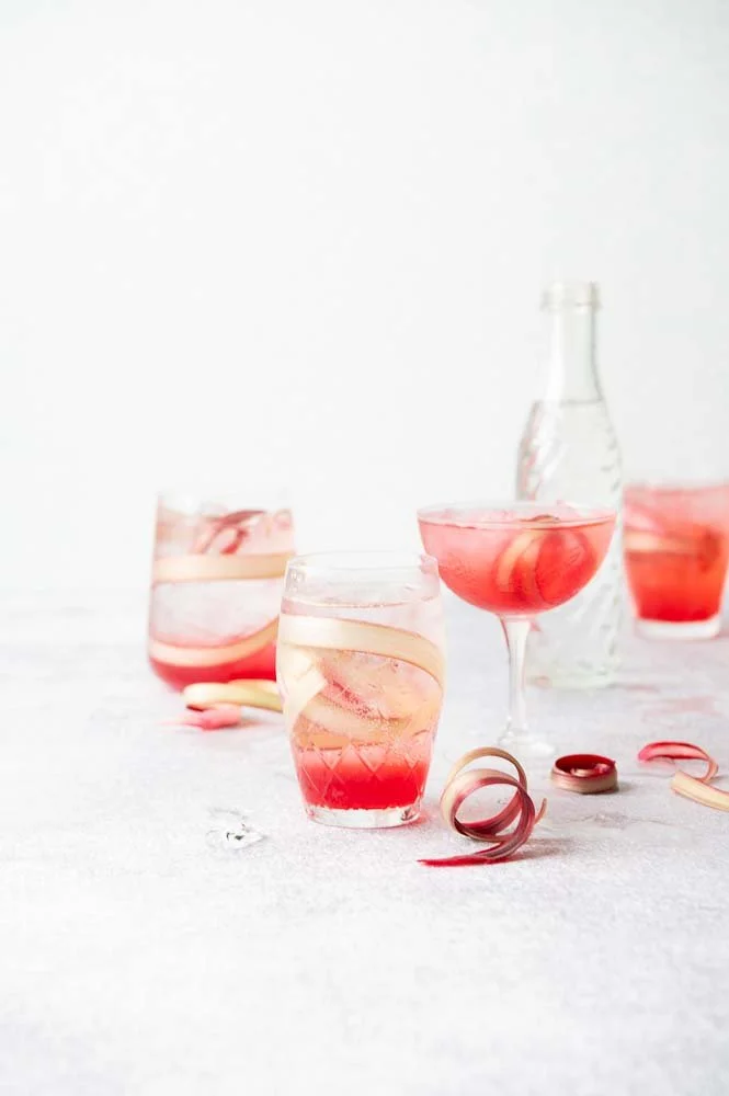 Four glasses with pink and orange layered drinks and a clear decanter on a light surface, with ribbons of rhubarb and ice cubes scattered around.